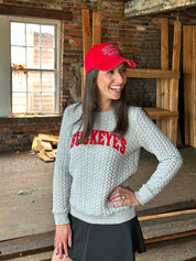 A woman wearing a light gray cable-knit sweater with 'HOCKEYES' in red across the chest, a red baseball cap, and a black skirt, standing in a rustic brick warehouse setting.
