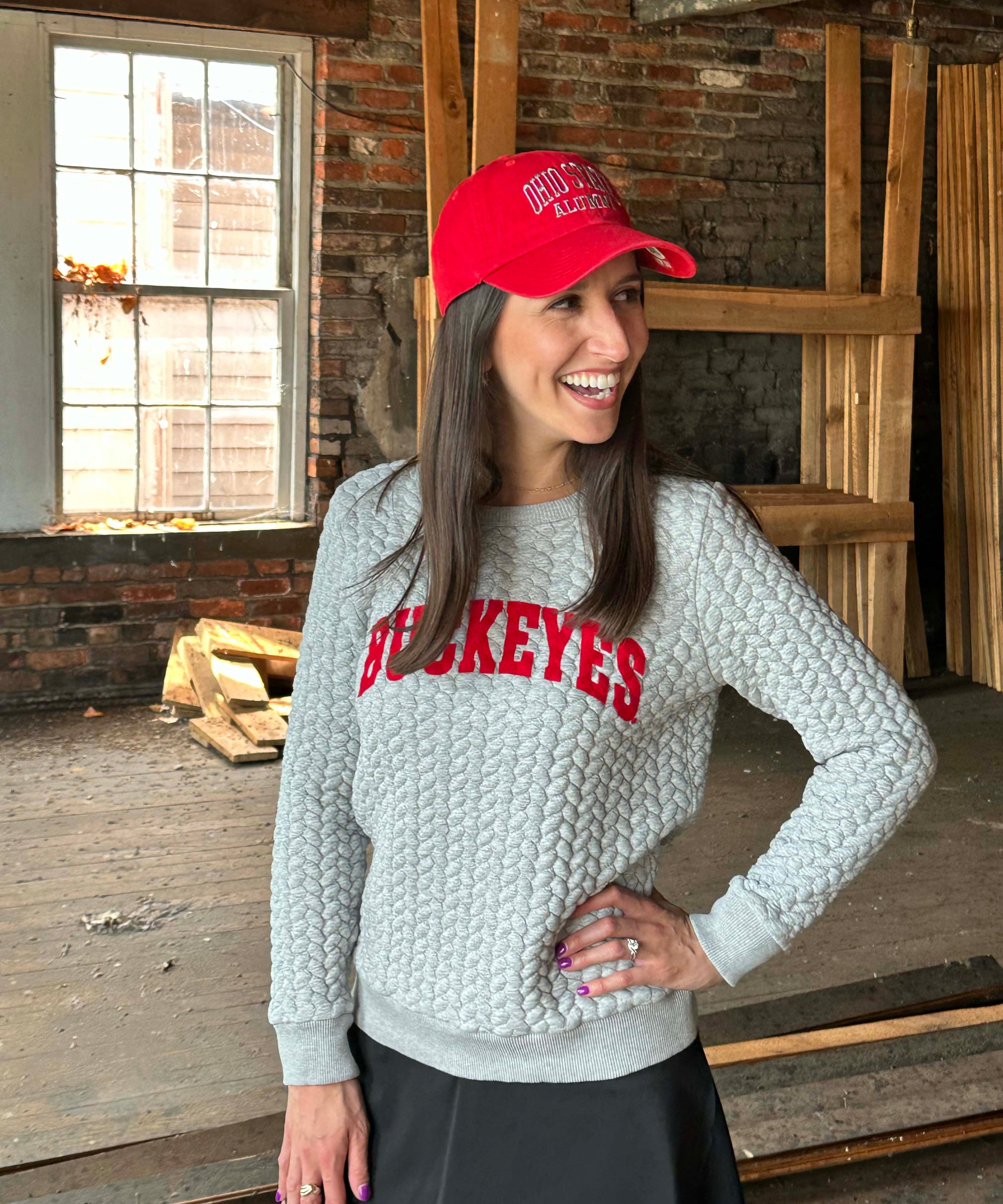 A woman wearing a light gray cable-knit sweater with 'HOCKEYES' in red across the chest, a red baseball cap, and a black skirt, standing in a rustic brick warehouse setting.