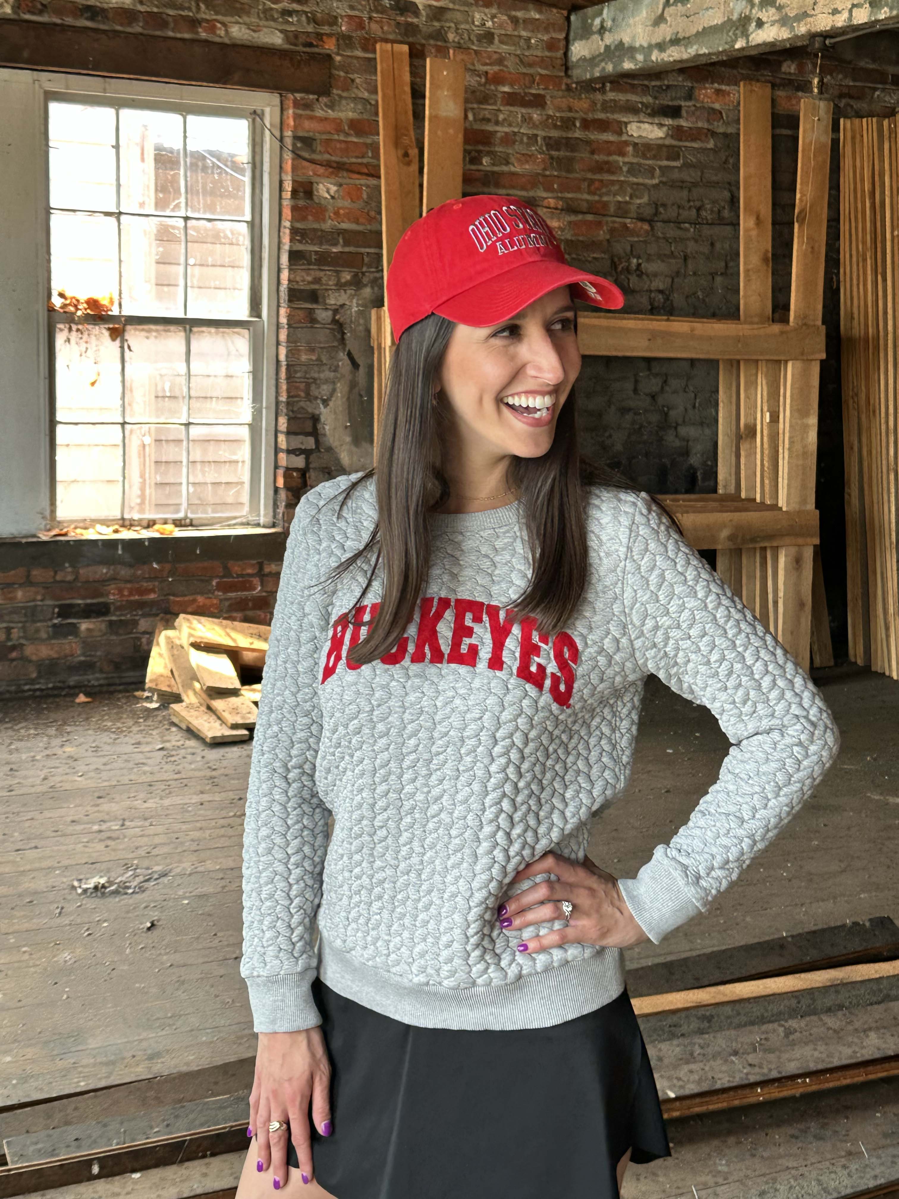 A woman wearing a light gray cable-knit sweater with 'HOCKEYES' in red across the chest, a red baseball cap, and a black skirt, standing in a rustic brick warehouse setting.