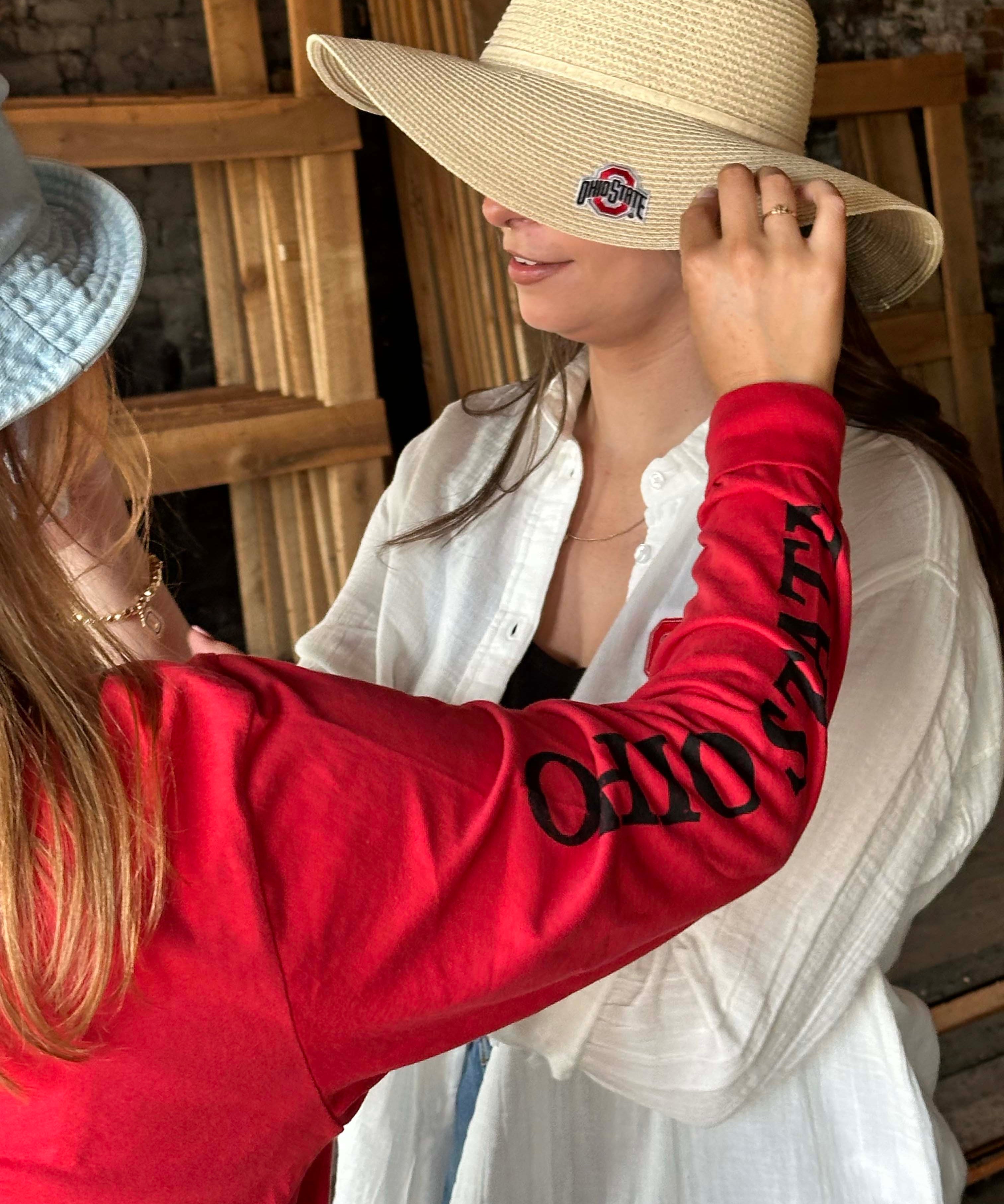 A woman wearing a large beige straw sun hat with a black and red Ohio State Buckeyes logo patch on the brim, while another person in a red sweatshirt adjusts the brim. The background shows a rustic wooden structure.