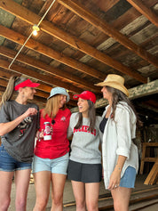 Four women standing together in a rustic wooden structure, wearing casual summer clothes including red visors, a red t-shirt, a gray sweatshirt with 'ROCKETS' text, denim shorts, and a straw hat.