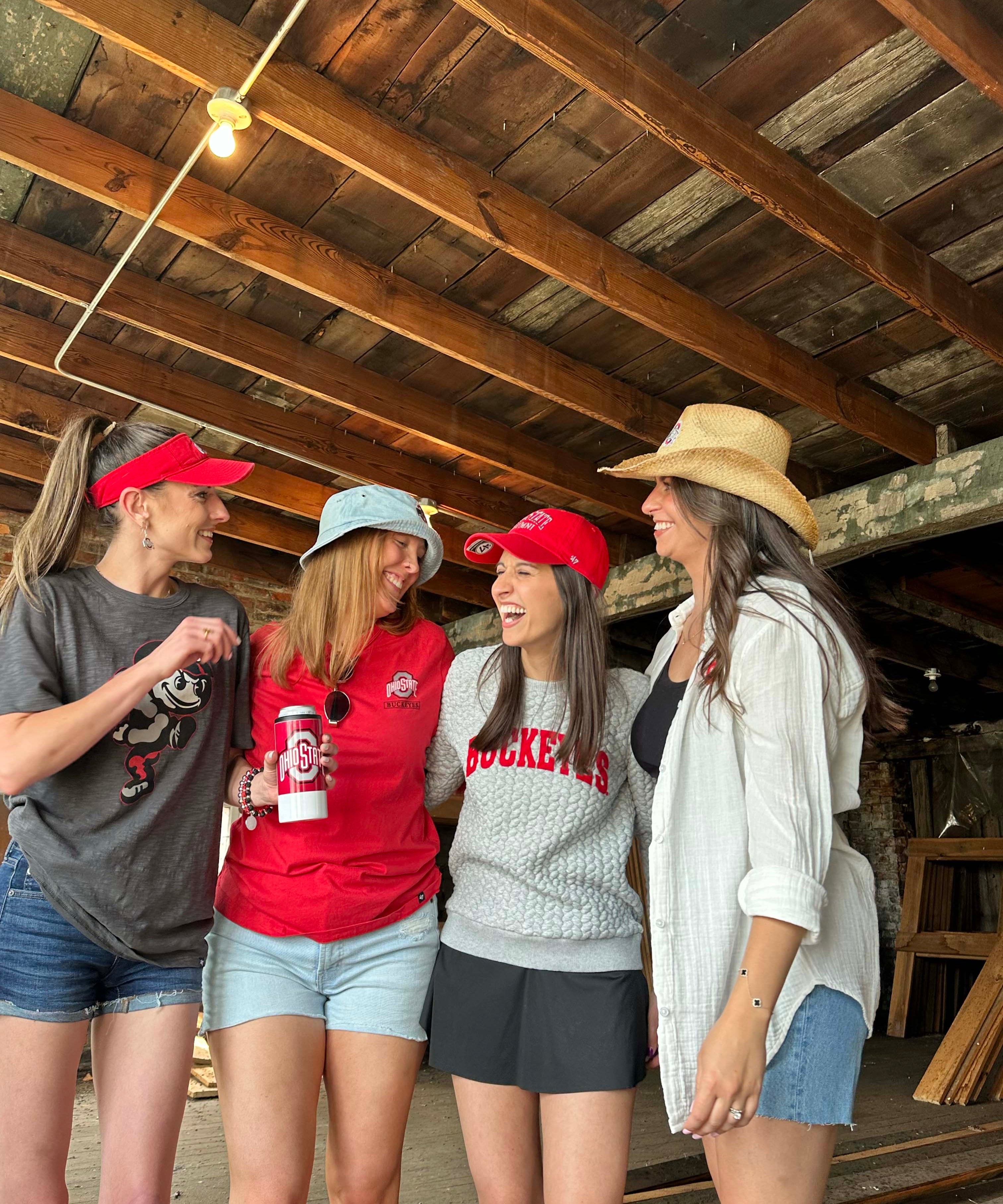 Four women standing together in a rustic wooden structure, wearing casual summer clothes including red visors, a red t-shirt, a gray sweatshirt with 'ROCKETS' text, denim shorts, and a straw hat.