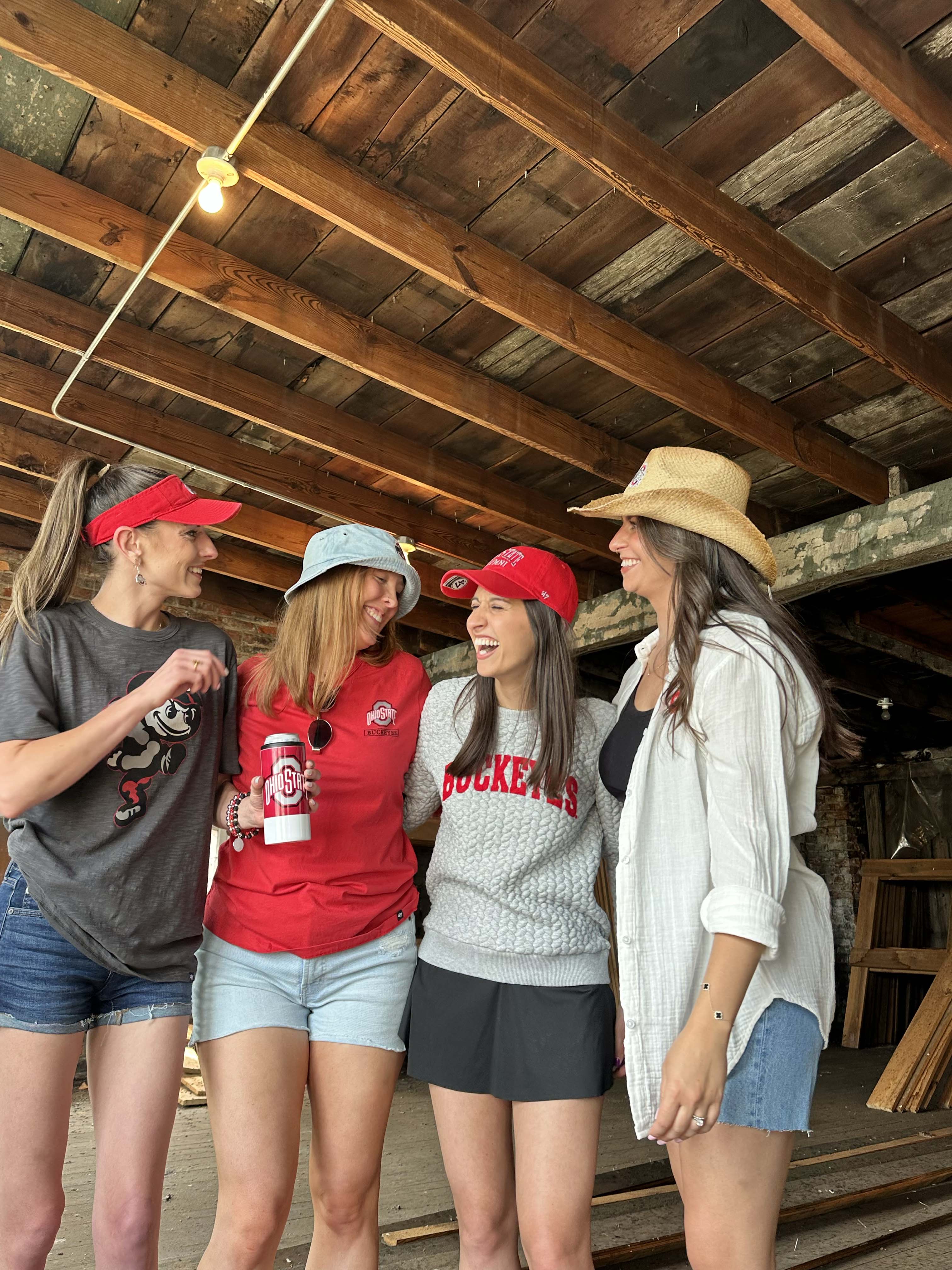 Four women standing together in a rustic wooden structure, wearing casual summer clothes including red visors, a red t-shirt, a gray sweatshirt with 'ROCKETS' text, denim shorts, and a straw hat.