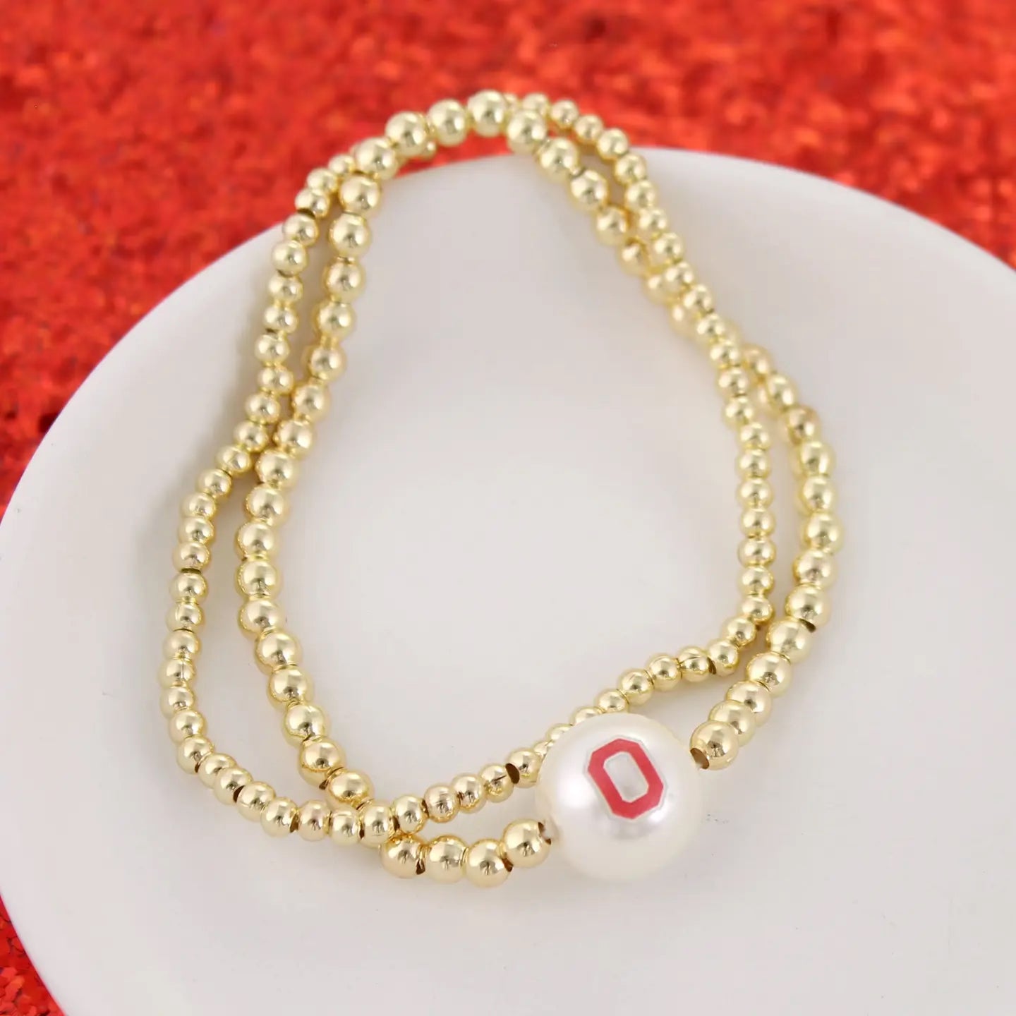 A double-strand beaded necklace with gold-toned beads and a white pearl bead featuring a red letter 'O' charm, displayed on a white plate against a textured red background.