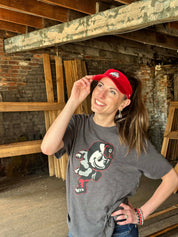 A woman posing in a rustic warehouse setting, wearing a gray graphic t-shirt with a cartoon character and a red visor.