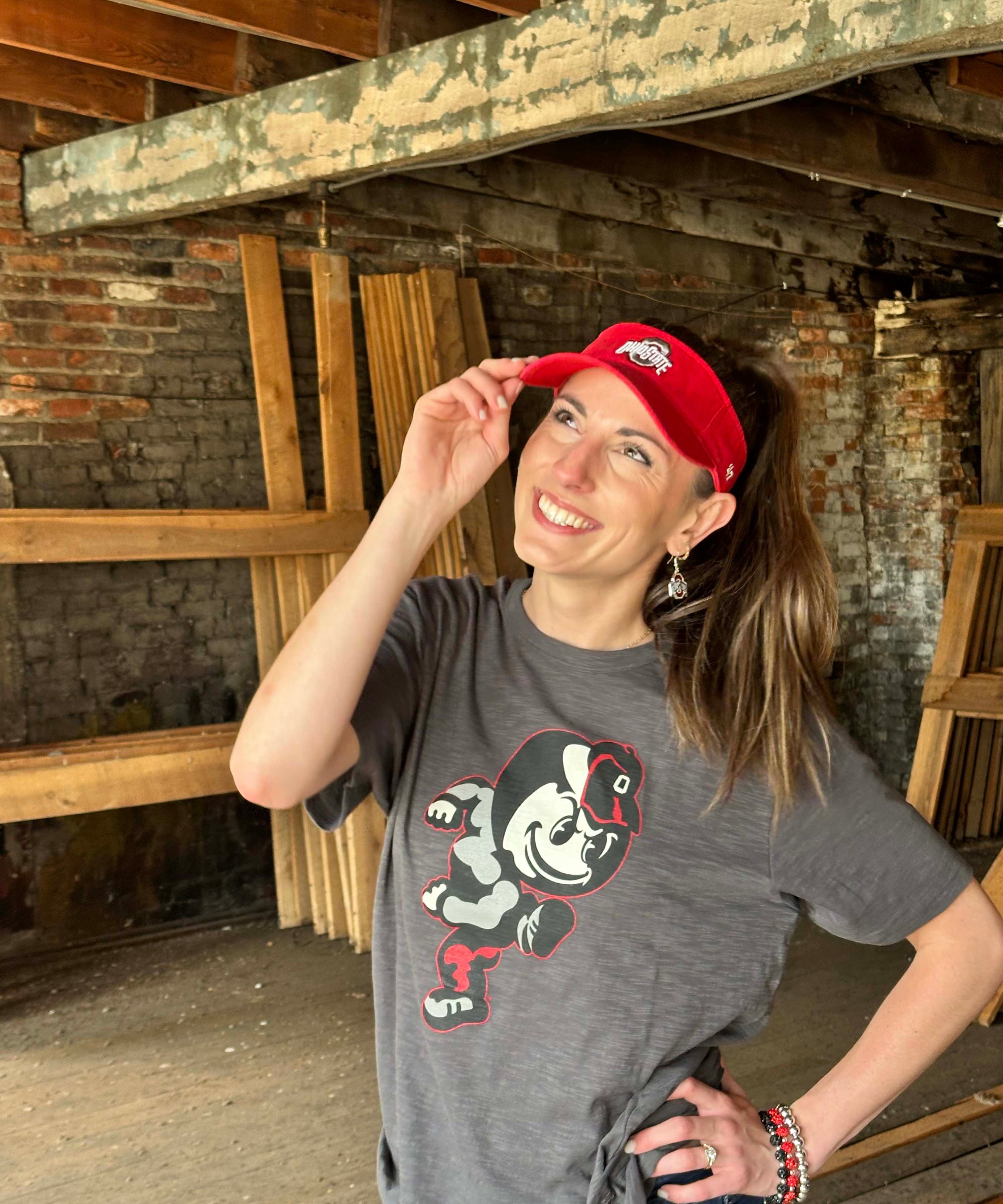 A woman posing in a rustic warehouse setting, wearing a gray graphic t-shirt with a cartoon character and a red visor.