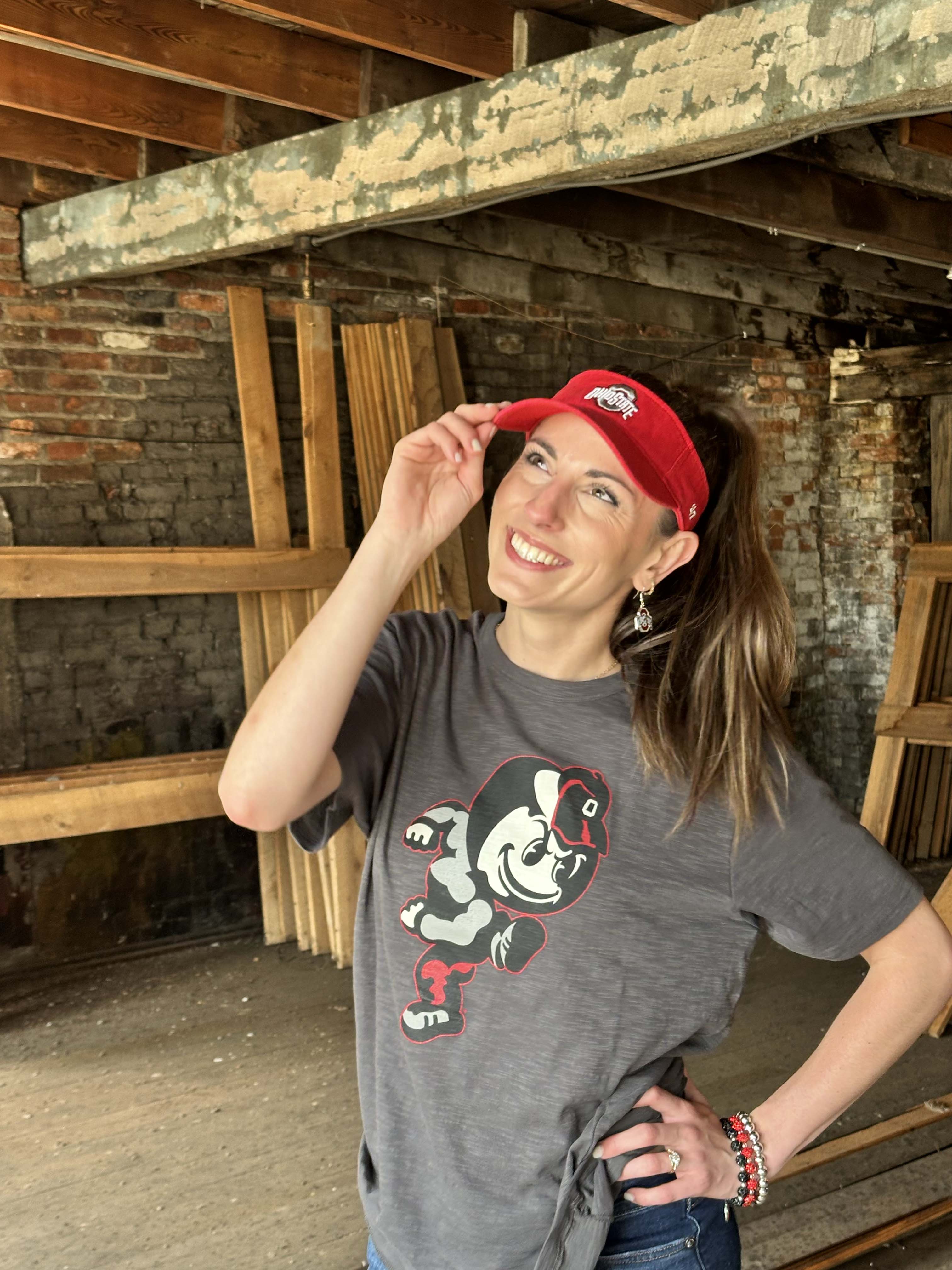 A woman posing in a rustic warehouse setting, wearing a gray graphic t-shirt with a cartoon character and a red visor.