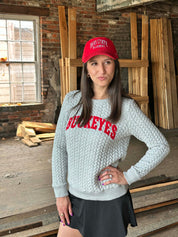 A woman wearing a light gray cable-knit sweatshirt with 'BUCKEYES' in red across the chest, a red baseball cap with 'OHIO STATE RAWDENS' in white, and a black skirt, standing in a rustic wood-paneled room.