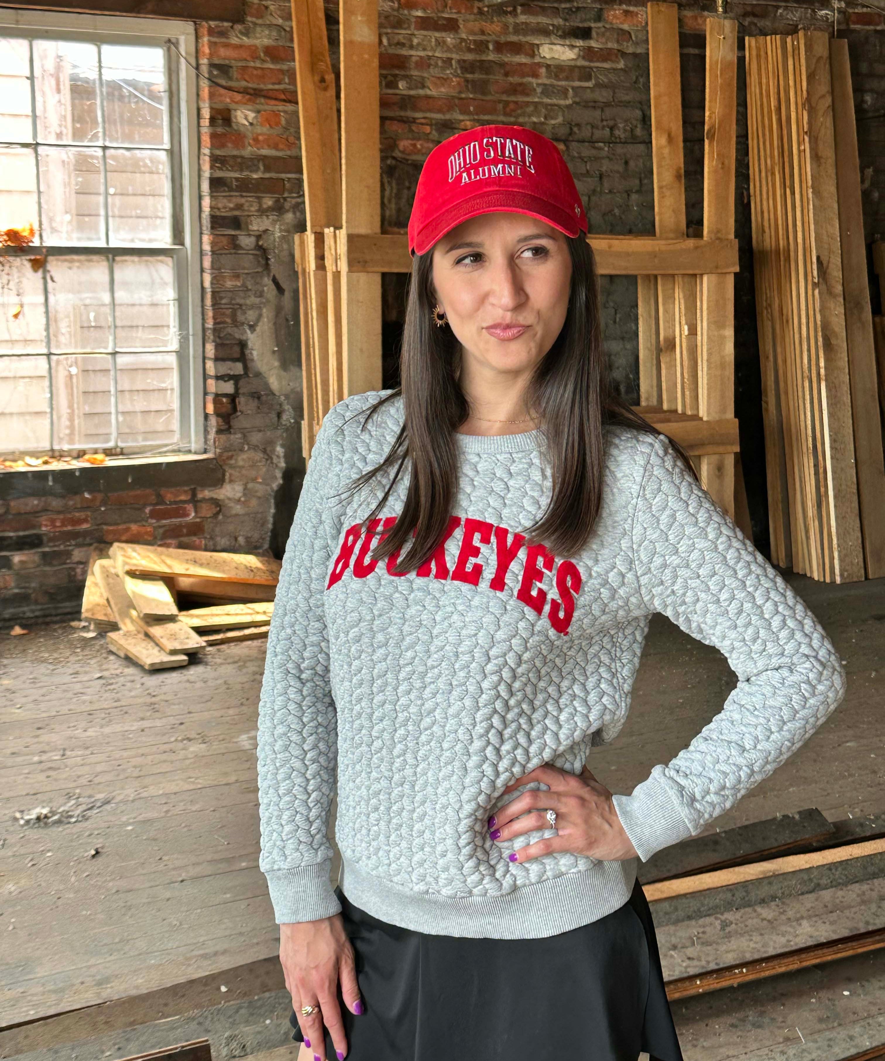 A woman wearing a light gray cable-knit sweatshirt with 'BUCKEYES' in red across the chest, a red baseball cap with 'OHIO STATE RAWDENS' in white, and a black skirt, standing in a rustic wood-paneled room.