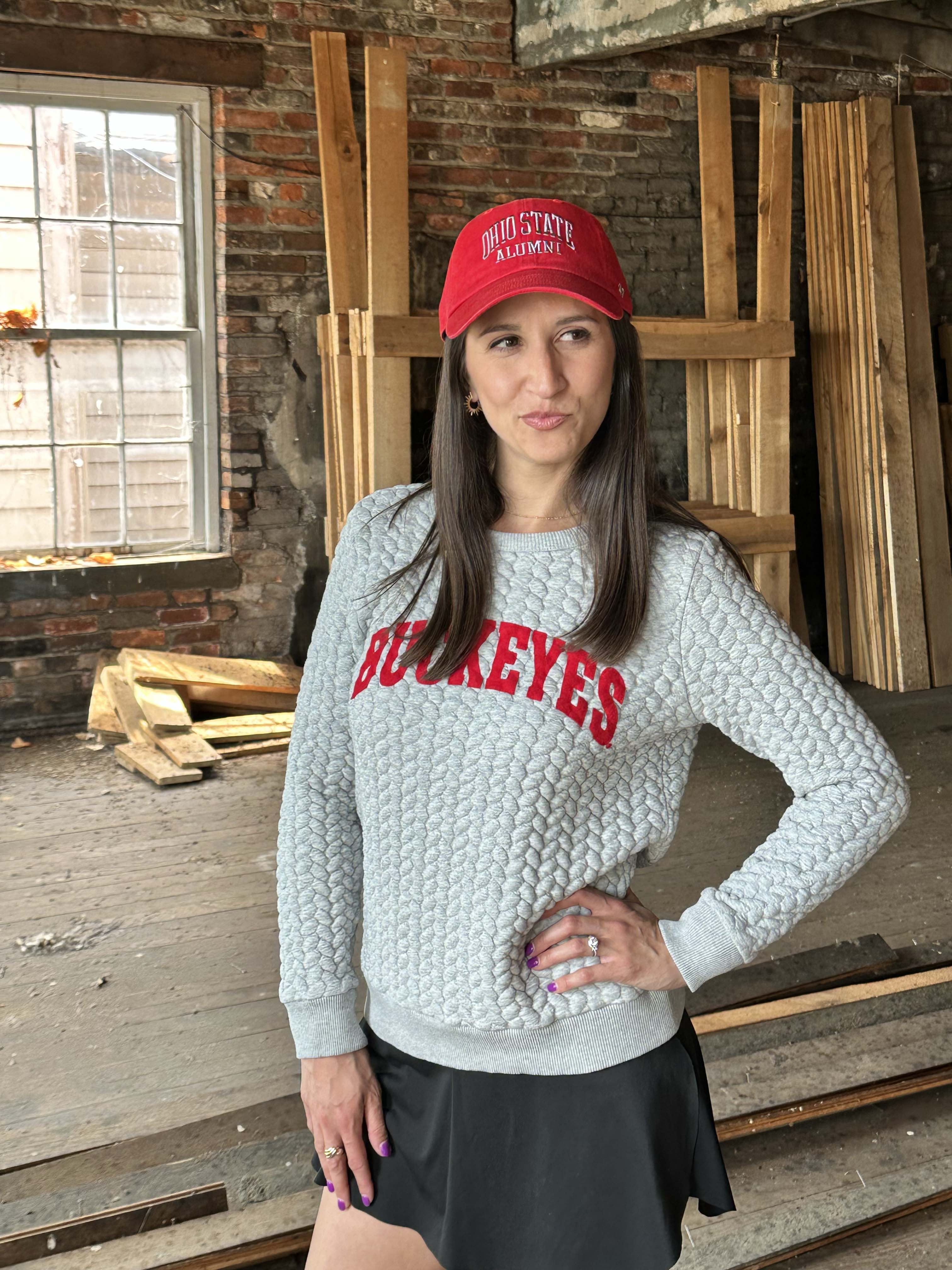 A woman wearing a light gray cable-knit sweatshirt with 'BUCKEYES' in red across the chest, a red baseball cap with 'OHIO STATE RAWDENS' in white, and a black skirt, standing in a rustic wood-paneled room.