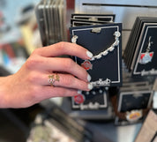 A hand with white nail polish holds a silver beaded bracelet with a red and white Ohio State Buckeyes logo charm on a black display card. The background shows a retail store display with other jewelry cards.