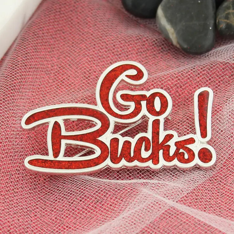 A red glittery enamel pin with the text 'Go Bucks!' in white cursive lettering, displayed on a red mesh fabric background with dark stones in the corner.
