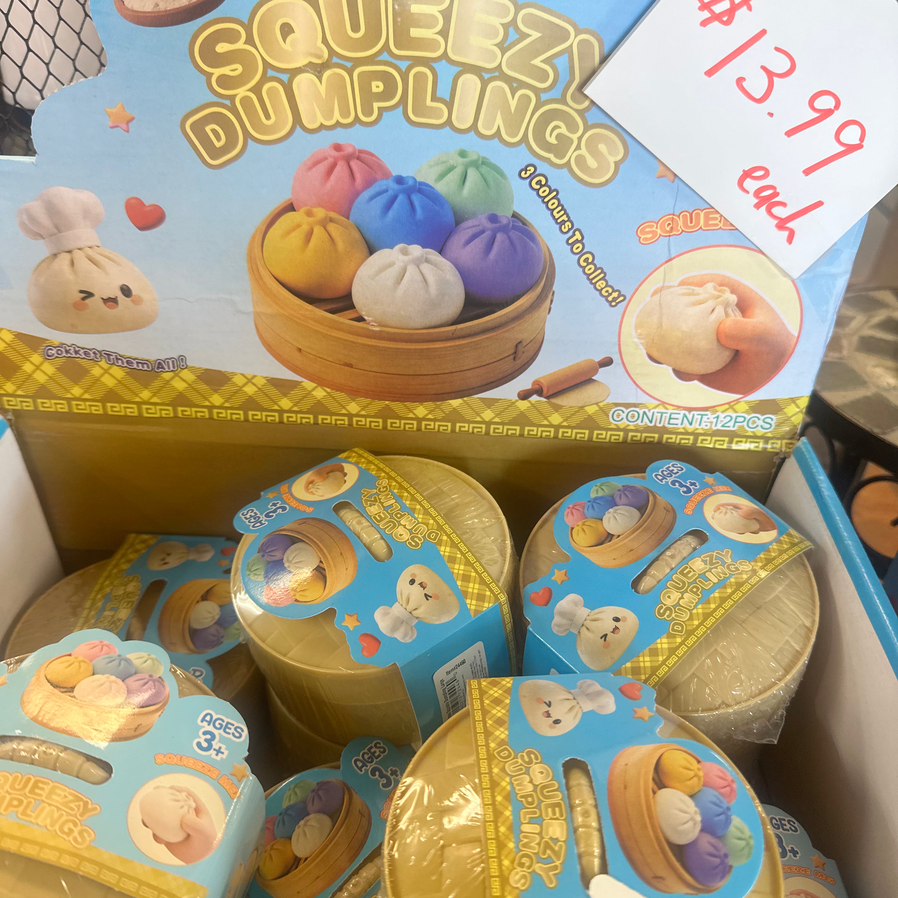 A retail display box filled with small round plastic toy dumpling containers. The top card reads 'SQUEEZI DUMPLINGS' with cartoon dumplings and a hand squeezing one, plus a handwritten price tag showing $13.