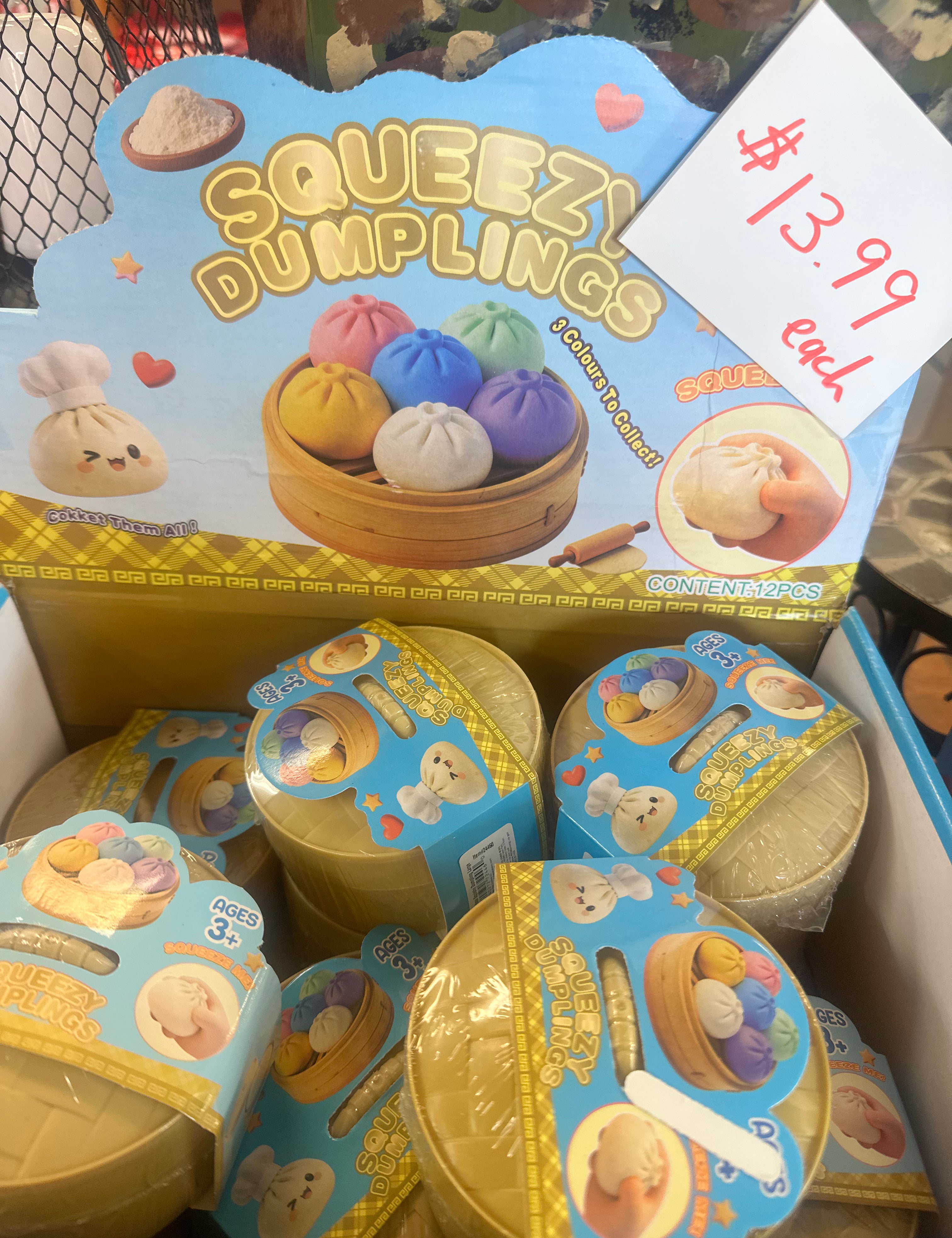 A retail display box filled with small round plastic toy dumpling containers. The top card reads 'SQUEEZI DUMPLINGS' with cartoon dumplings and a hand squeezing one, plus a handwritten price tag showing $13.