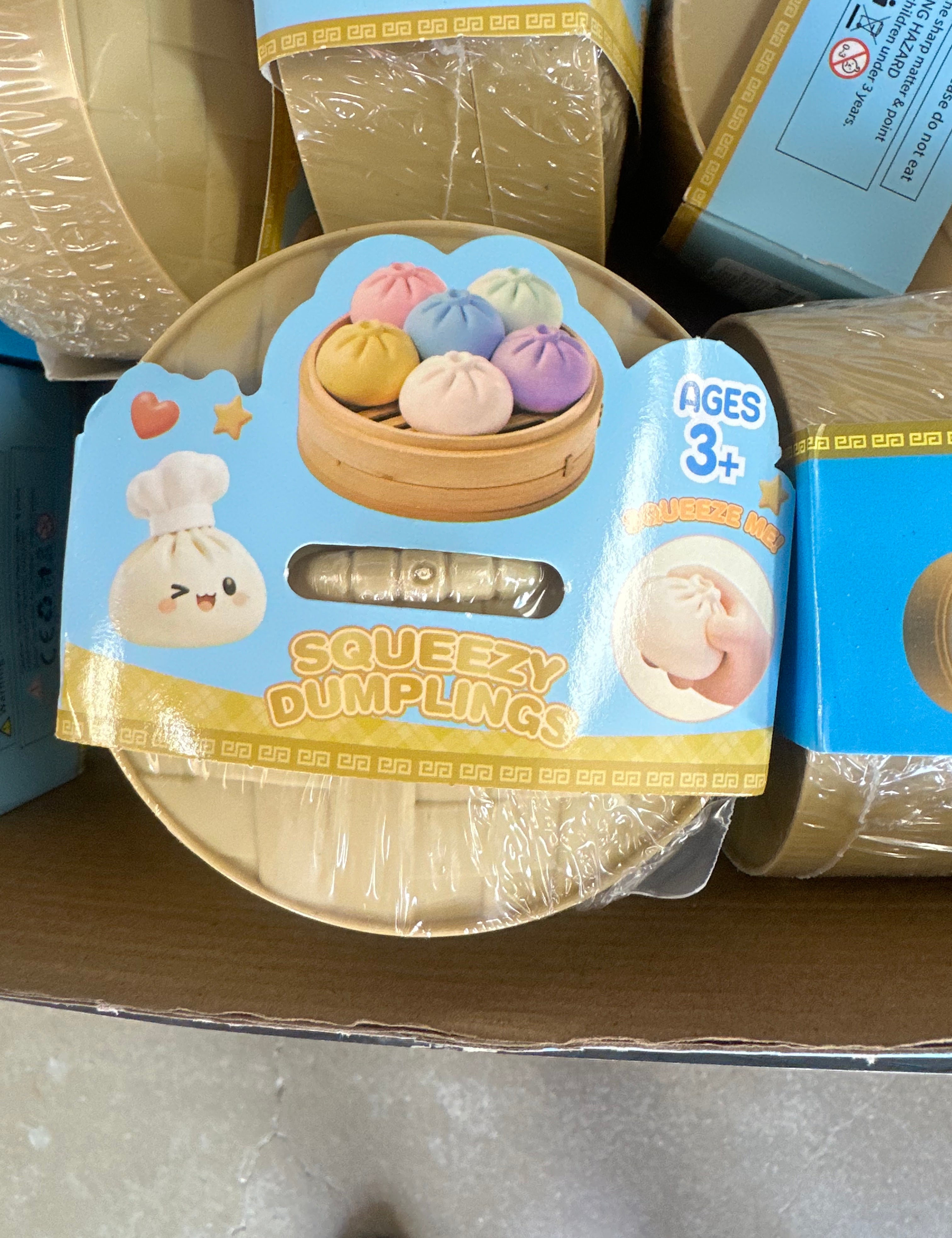 A round toy container labeled 'Squeezzy Dumplings' with a cartoon dumpling character and a photo of colorful dumplings in a steamer, shown on a cardboard shelf next to other similar packages.