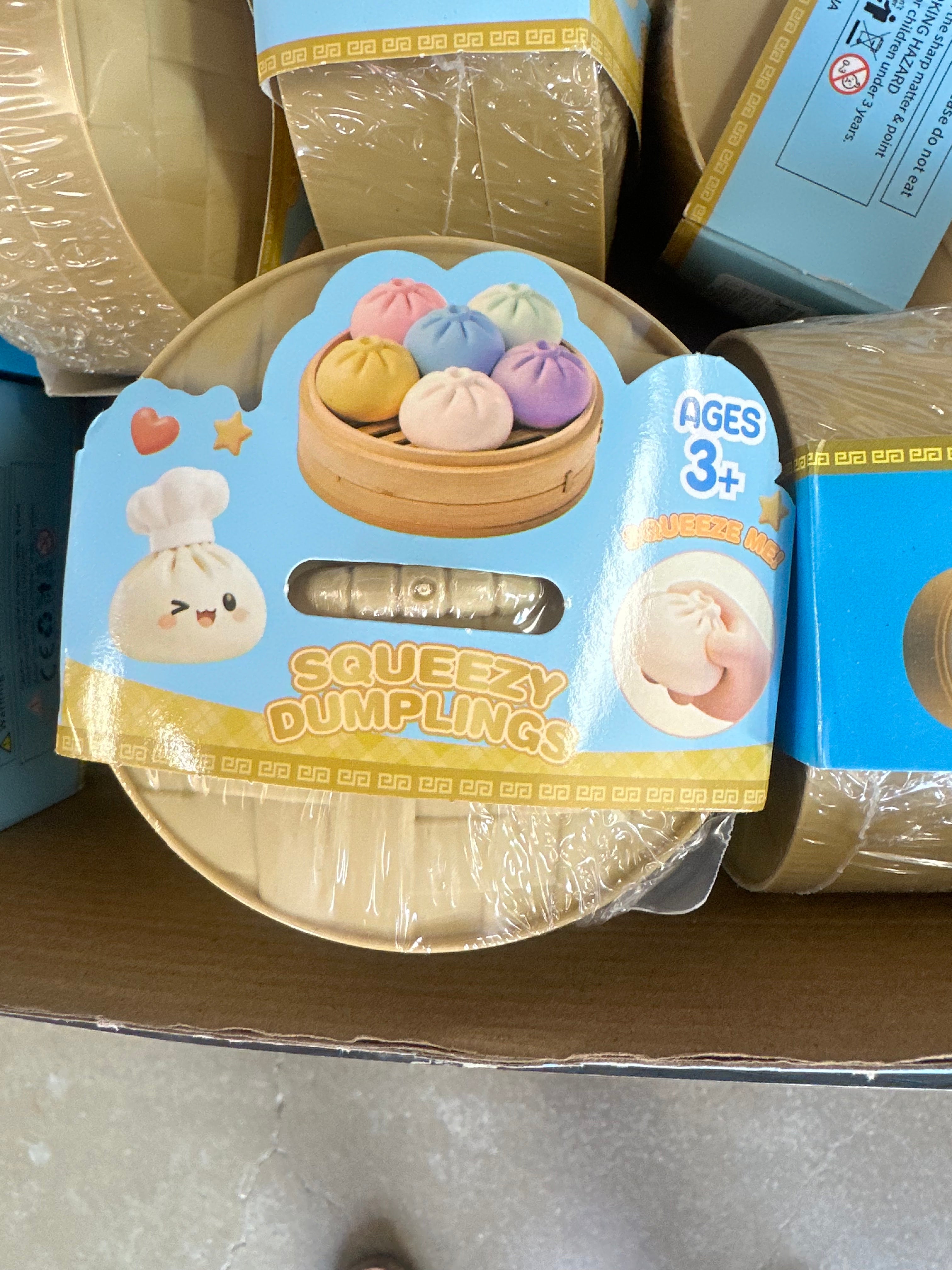 A round toy container labeled 'Squeezzy Dumplings' with a cartoon dumpling character and a photo of colorful dumplings in a steamer, shown on a cardboard shelf next to other similar packages.