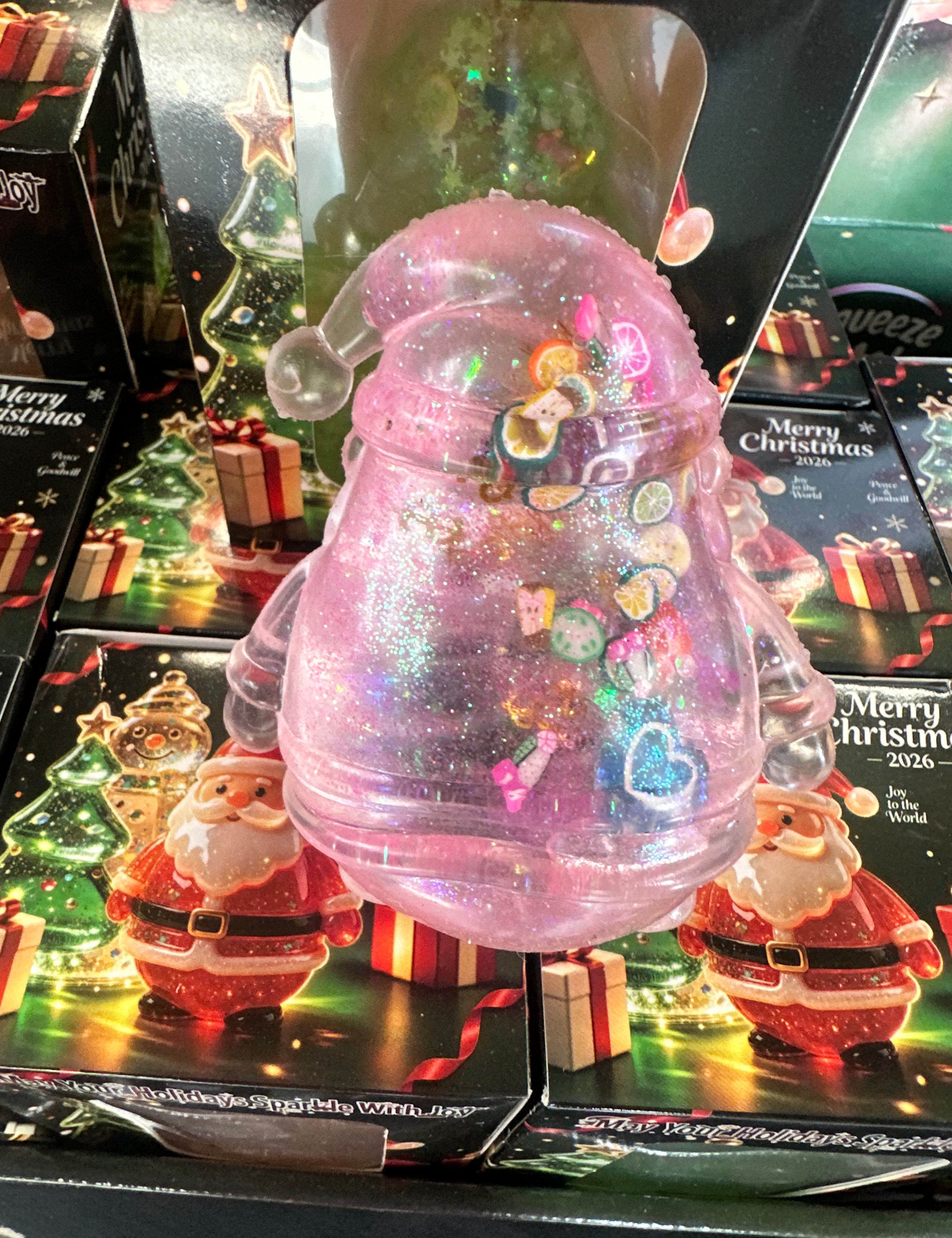 A pink glittery Christmas ornament-shaped container with a clear Santa hat and colorful heart-shaped embellishments, displayed in front of holiday-themed packaging featuring Santa Claus and 'Merry Christmas' designs.