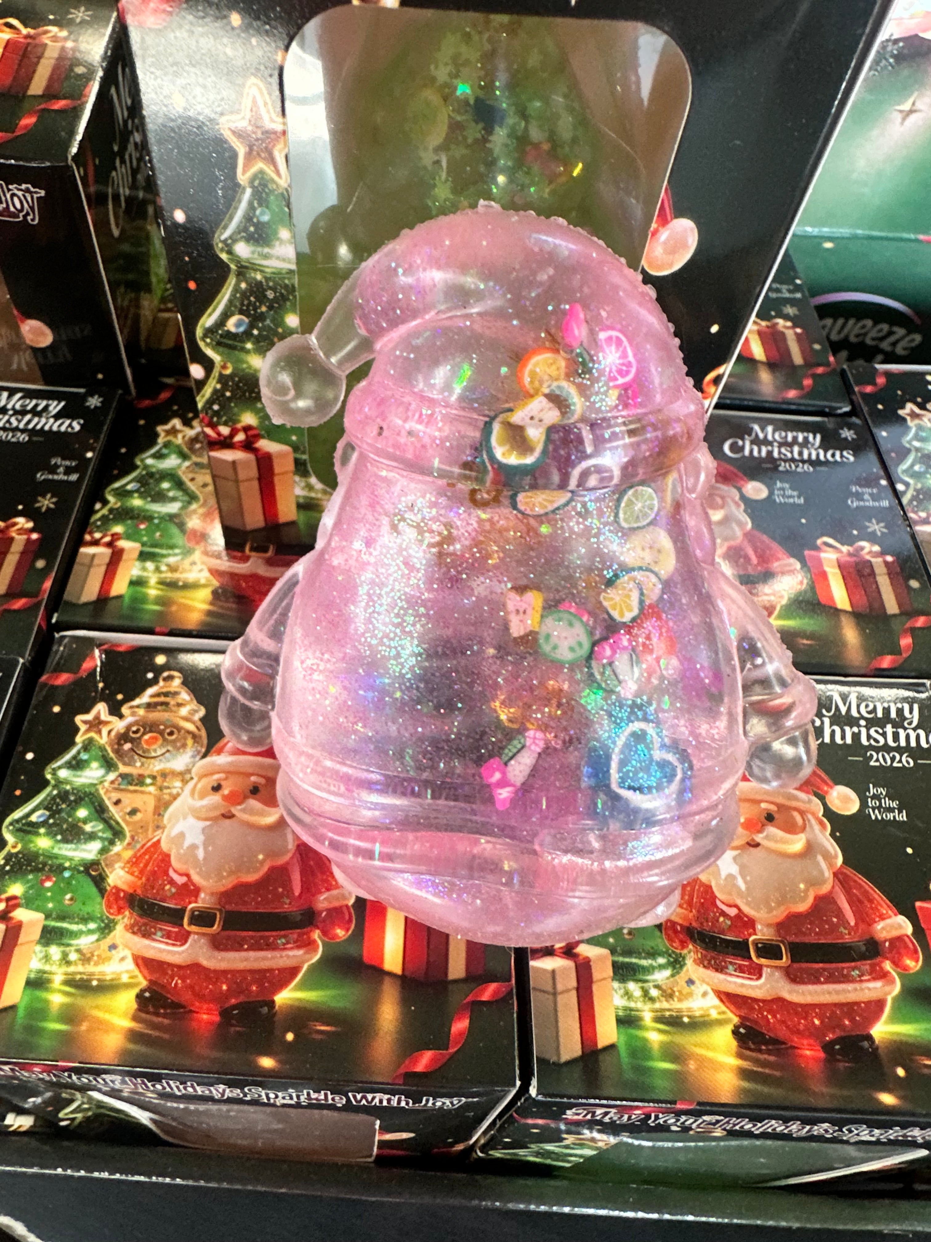 A pink glittery Christmas ornament-shaped container with a clear Santa hat and colorful heart-shaped embellishments, displayed in front of holiday-themed packaging featuring Santa Claus and 'Merry Christmas' designs.
