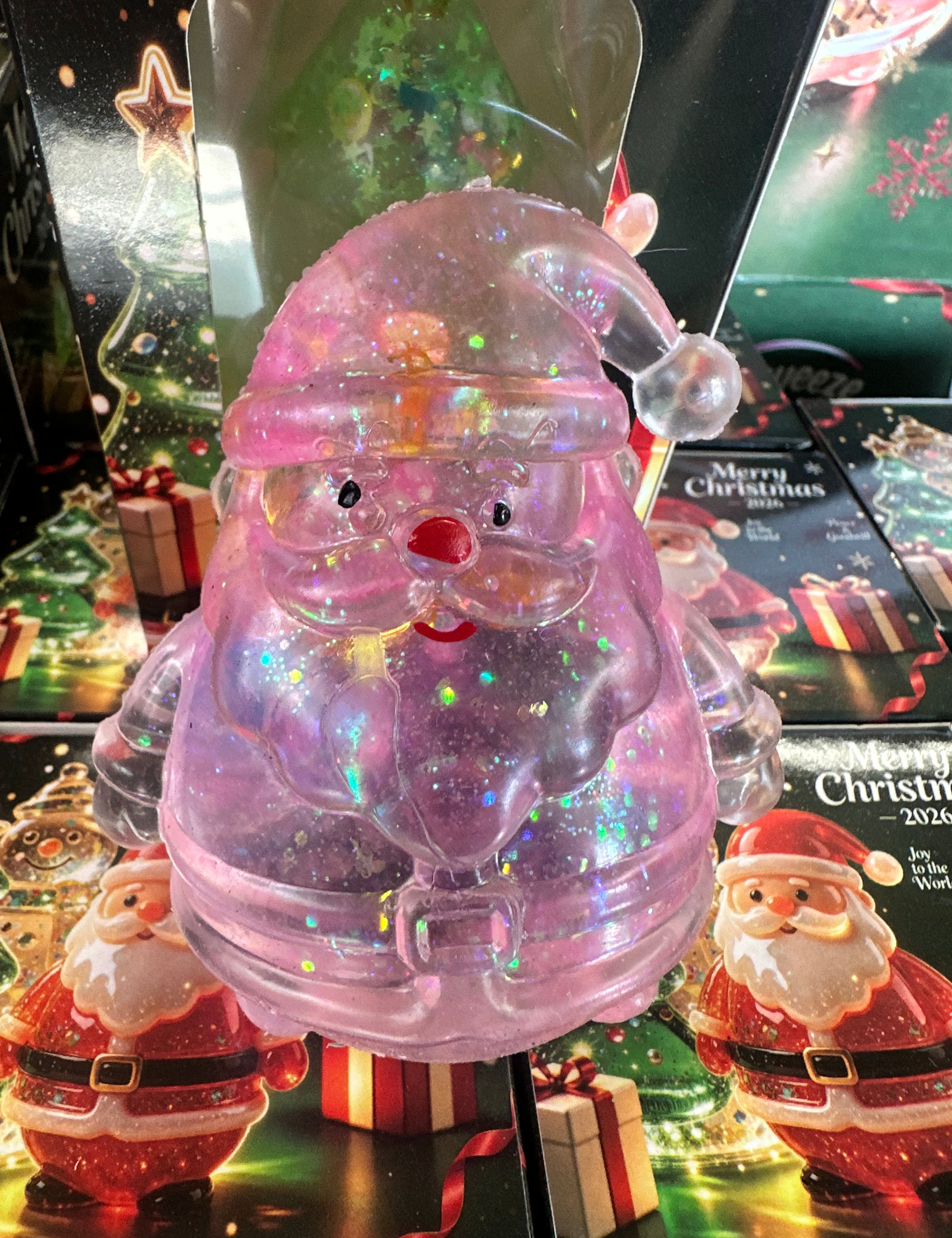 A translucent pink Santa Claus figurine filled with iridescent glitter, displayed in front of Christmas-themed packaging and other Santa decorations.