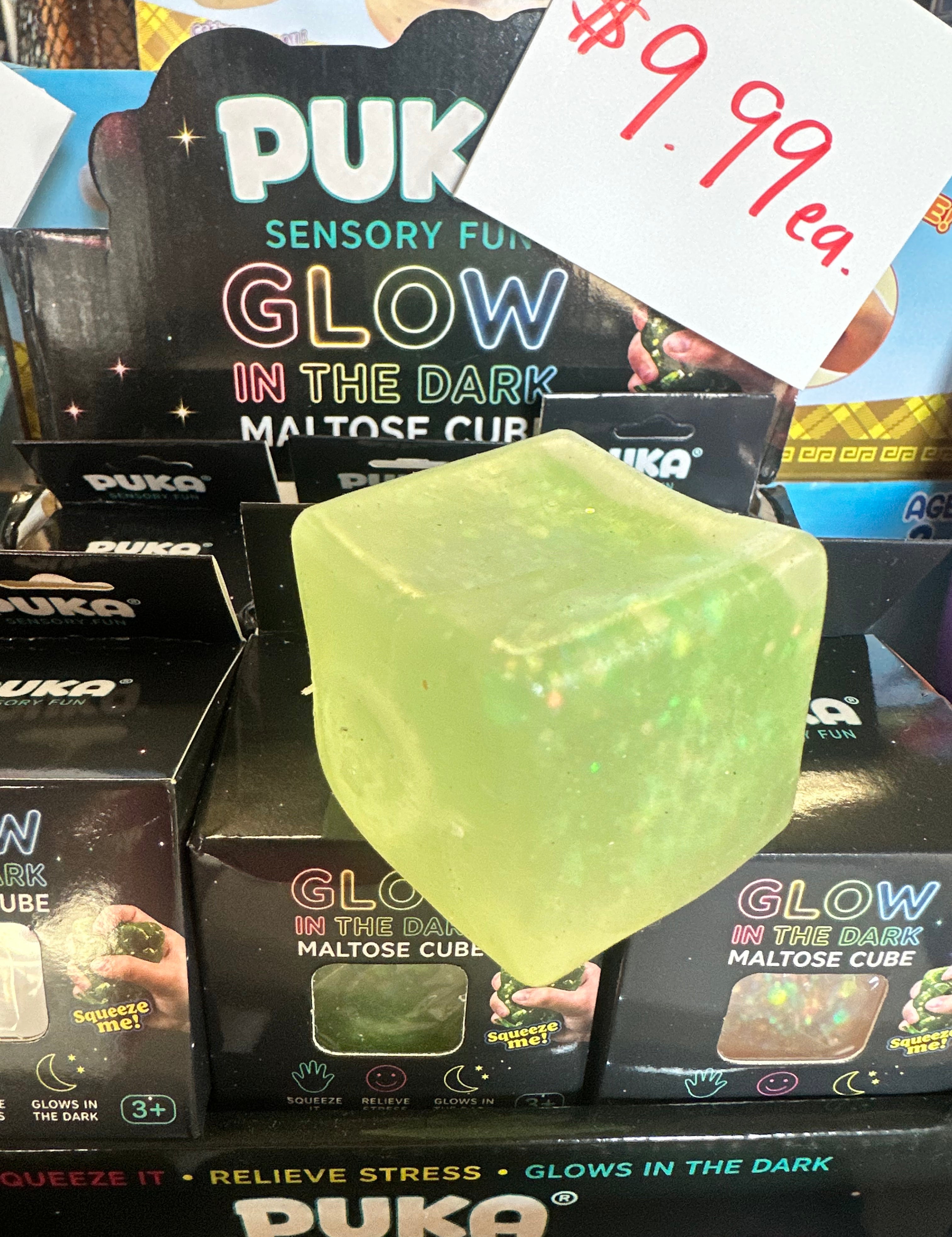 A translucent pale green glow-in-the-dark maltose cube toy with embedded glitter, displayed in front of its retail packaging. A handwritten price tag reading '$9.
