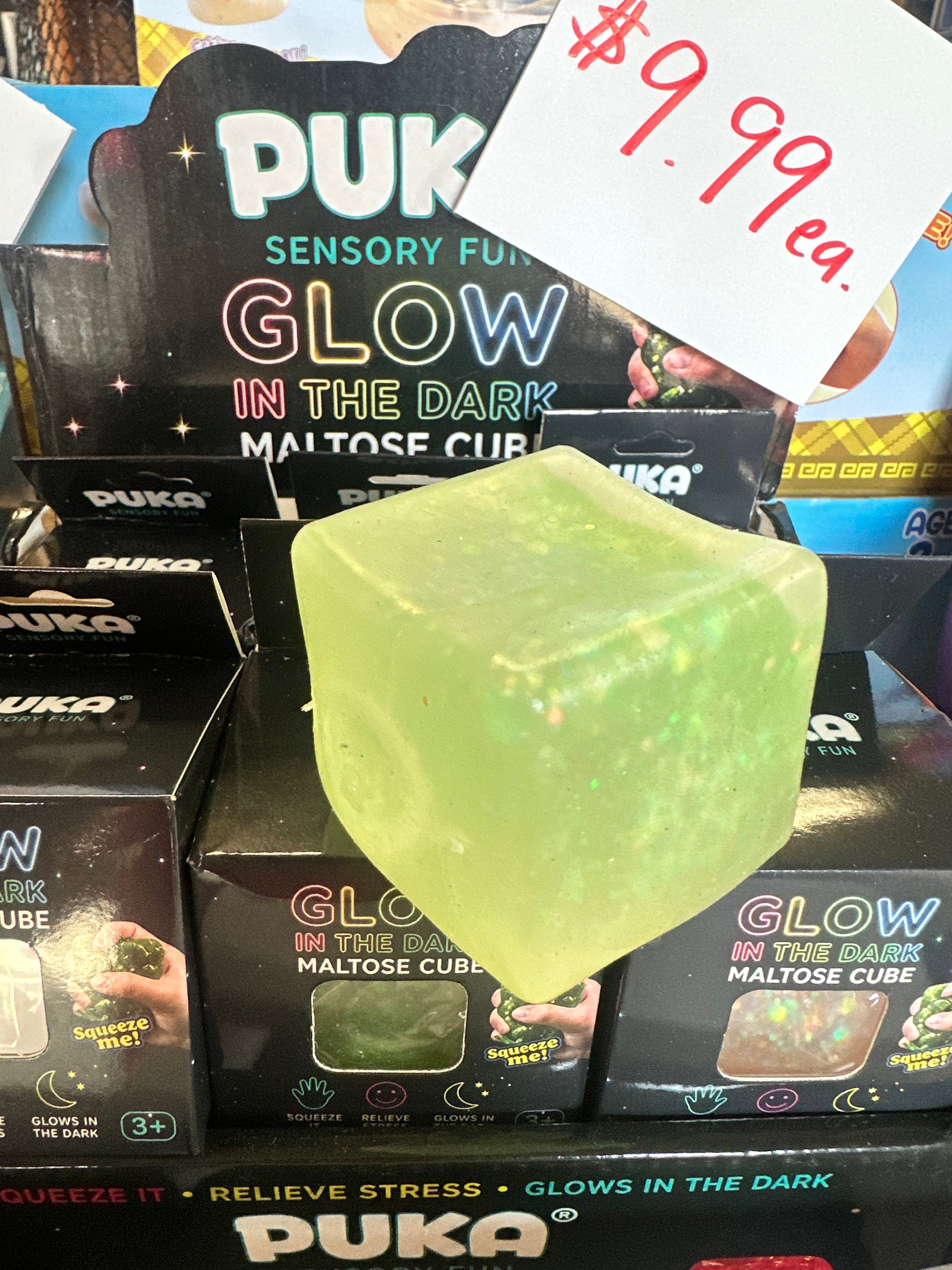 A translucent pale green glow-in-the-dark maltose cube toy with embedded glitter, displayed in front of its retail packaging. A handwritten price tag reading '$9.