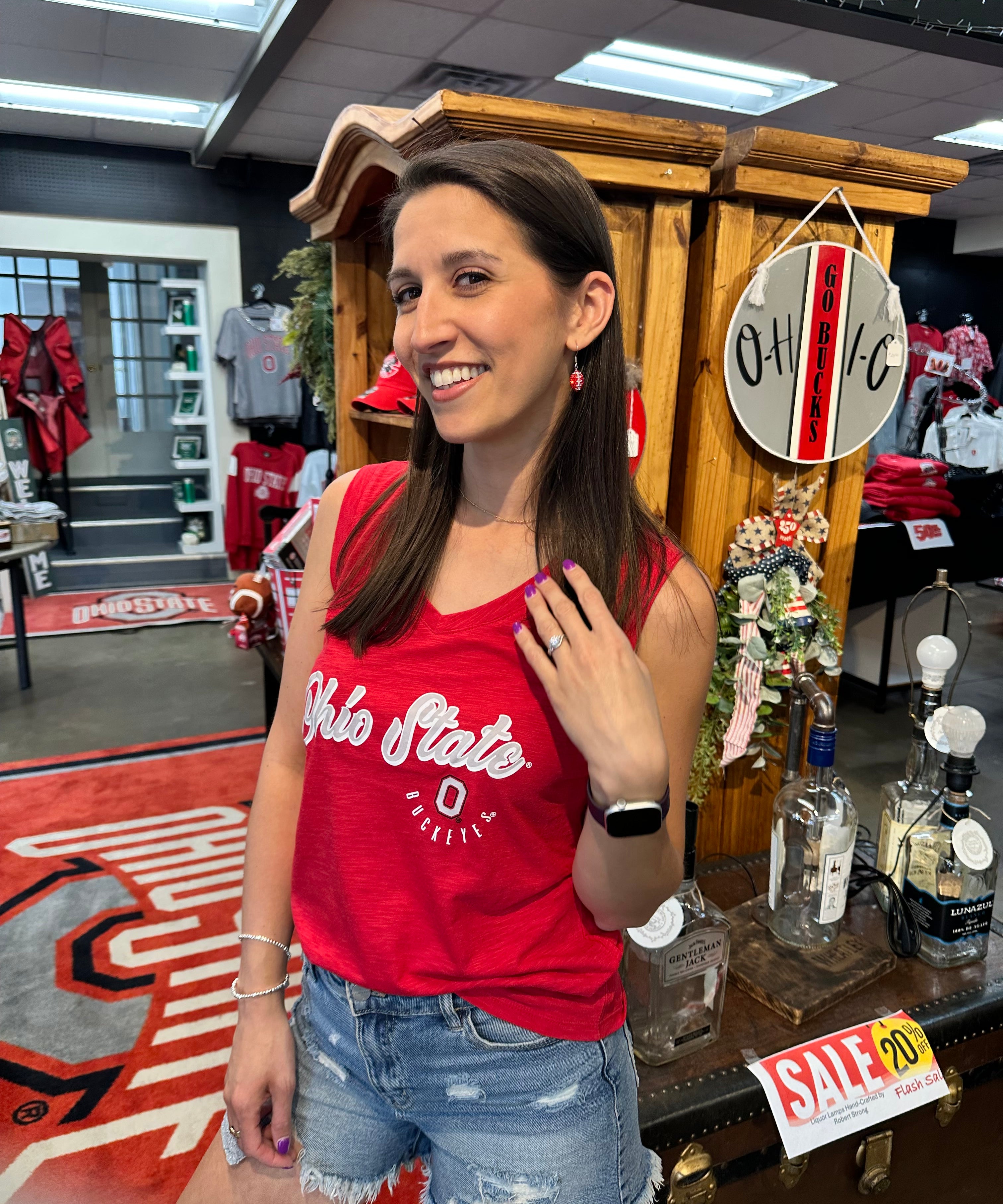 A woman smiling in a retail store wearing a red tank top with 'Ohio State' printed in white, paired with distressed denim shorts. The background shows a wooden display cabinet, a 'OHIO' sign, and a liquor display counter.