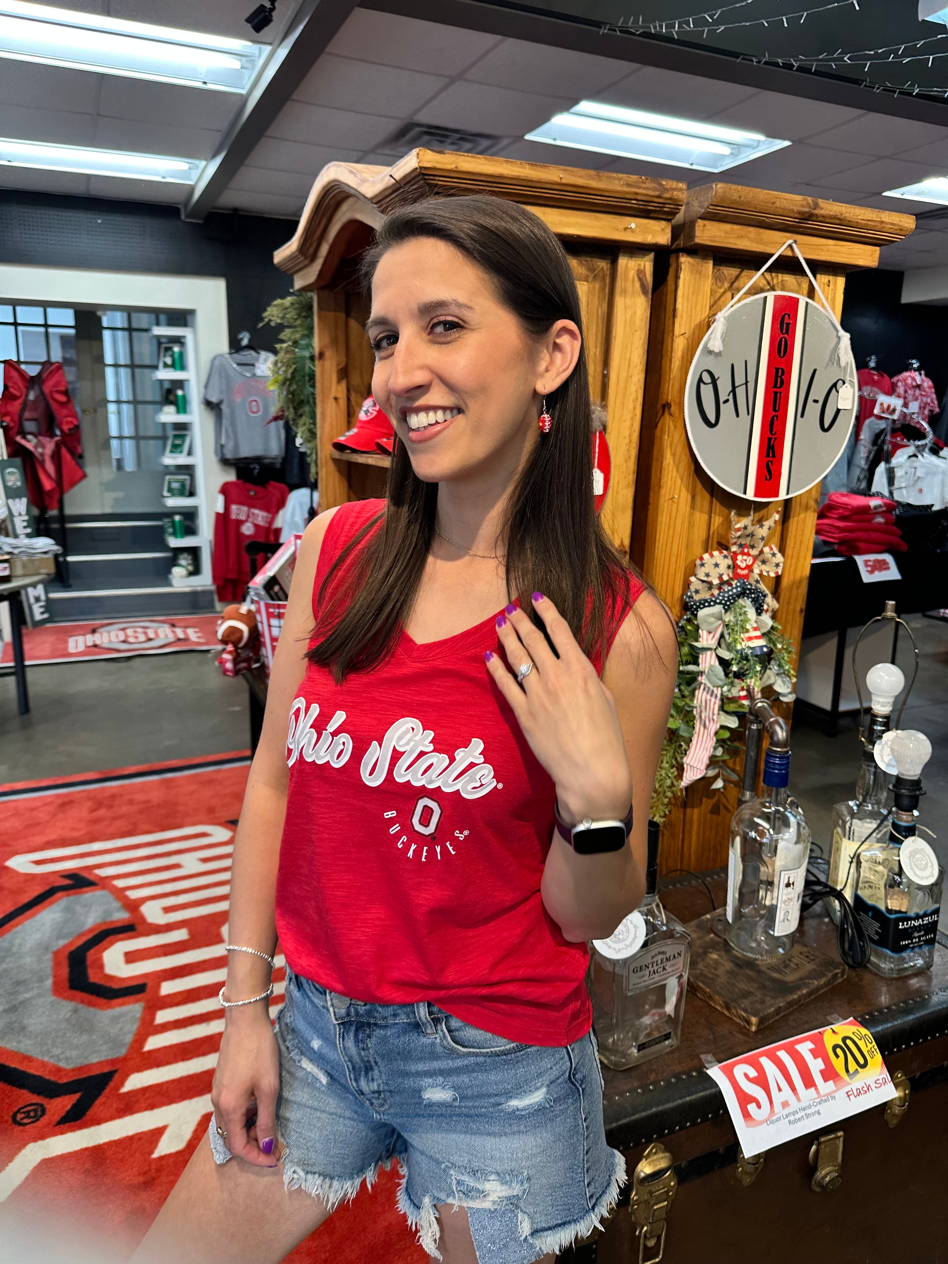 A woman smiling in a retail store wearing a red tank top with 'Ohio State' printed in white, paired with distressed denim shorts. The background shows a wooden display cabinet, a 'OHIO' sign, and a liquor display counter.