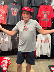 A man wearing an Ohio State Buckeyes gray performance t-shirt and a red bucket hat stands in a retail store with arms outstretched, surrounded by various Ohio State themed t-shirts hanging on a black pegboard.