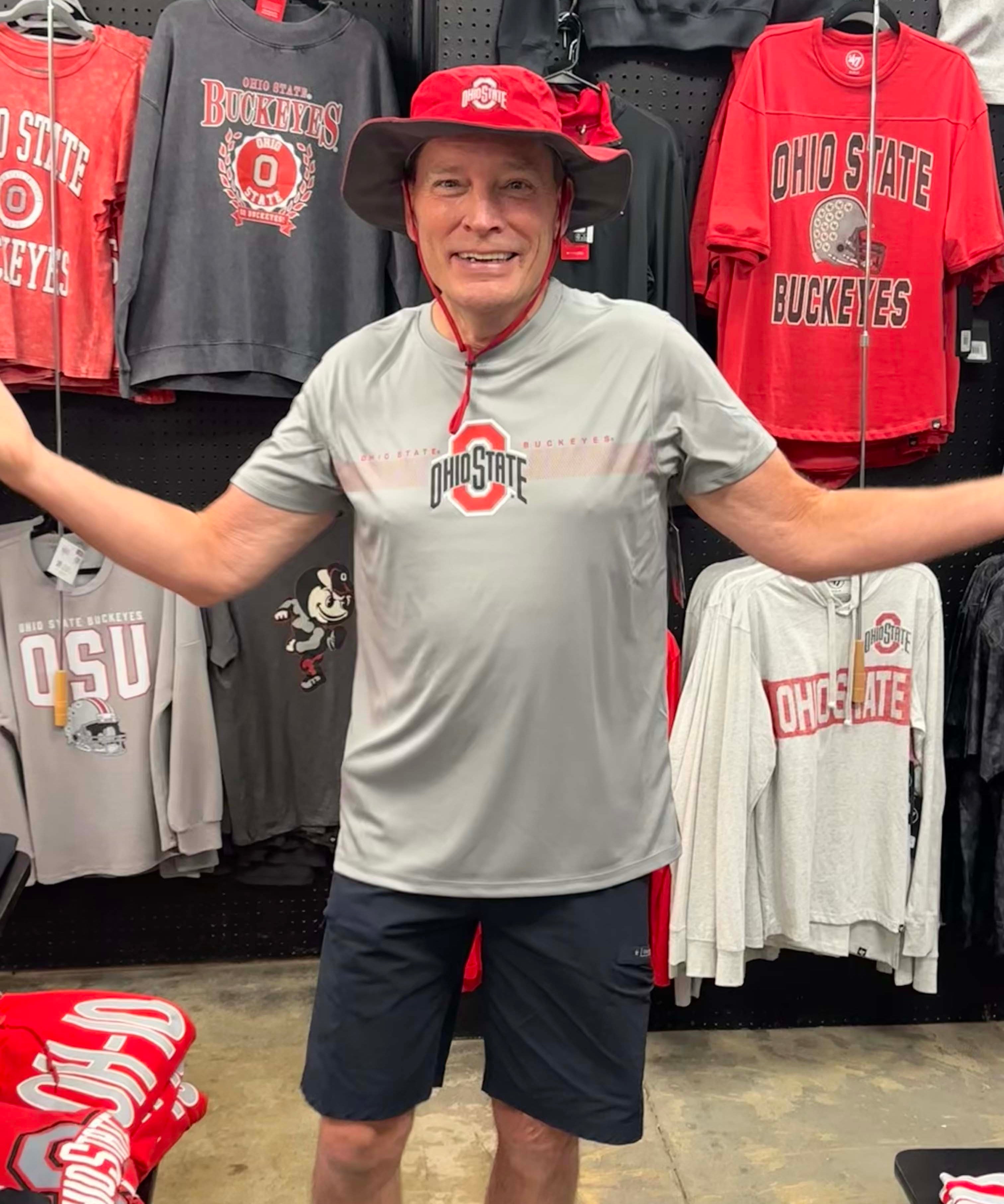 A man wearing an Ohio State Buckeyes gray performance t-shirt and a red bucket hat stands in a retail store with arms outstretched, surrounded by various Ohio State themed t-shirts hanging on a black pegboard.