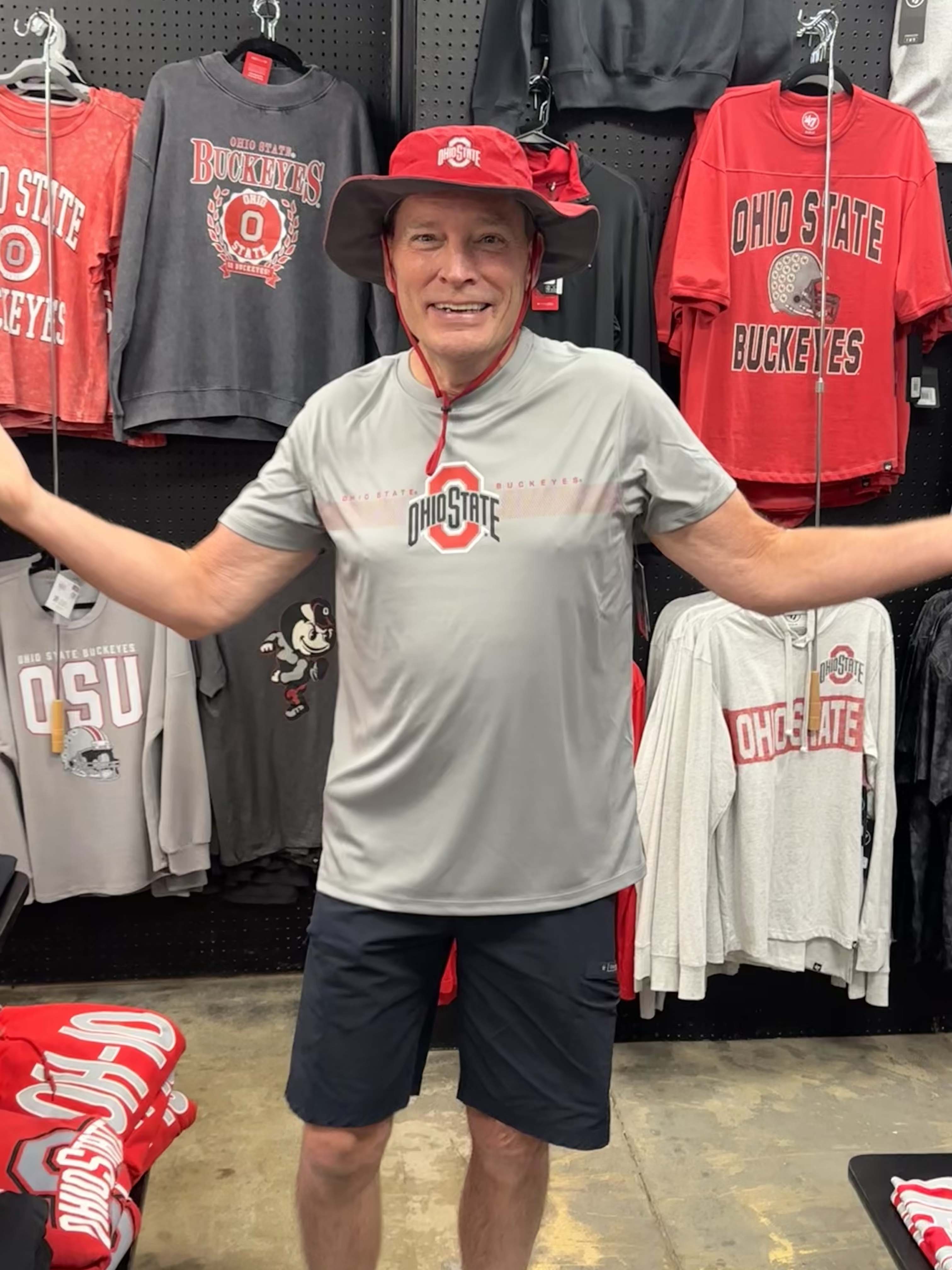 A man wearing an Ohio State Buckeyes gray performance t-shirt and a red bucket hat stands in a retail store with arms outstretched, surrounded by various Ohio State themed t-shirts hanging on a black pegboard.