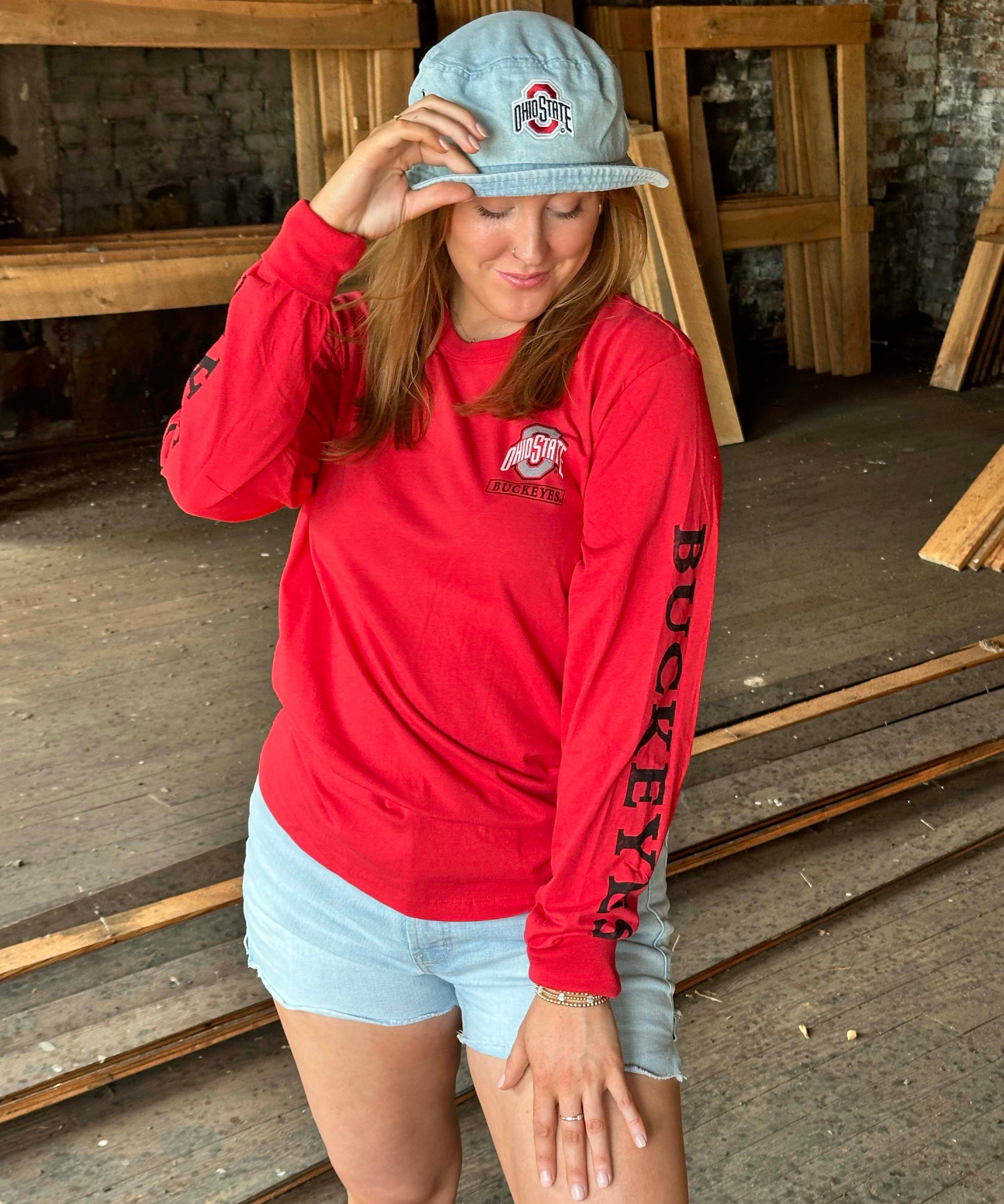 A woman wearing a bright red long-sleeve Ohio State Buckeyes sweatshirt, light blue denim shorts, and a light blue baseball cap with the Ohio State logo, standing in a rustic wood-paneled room.