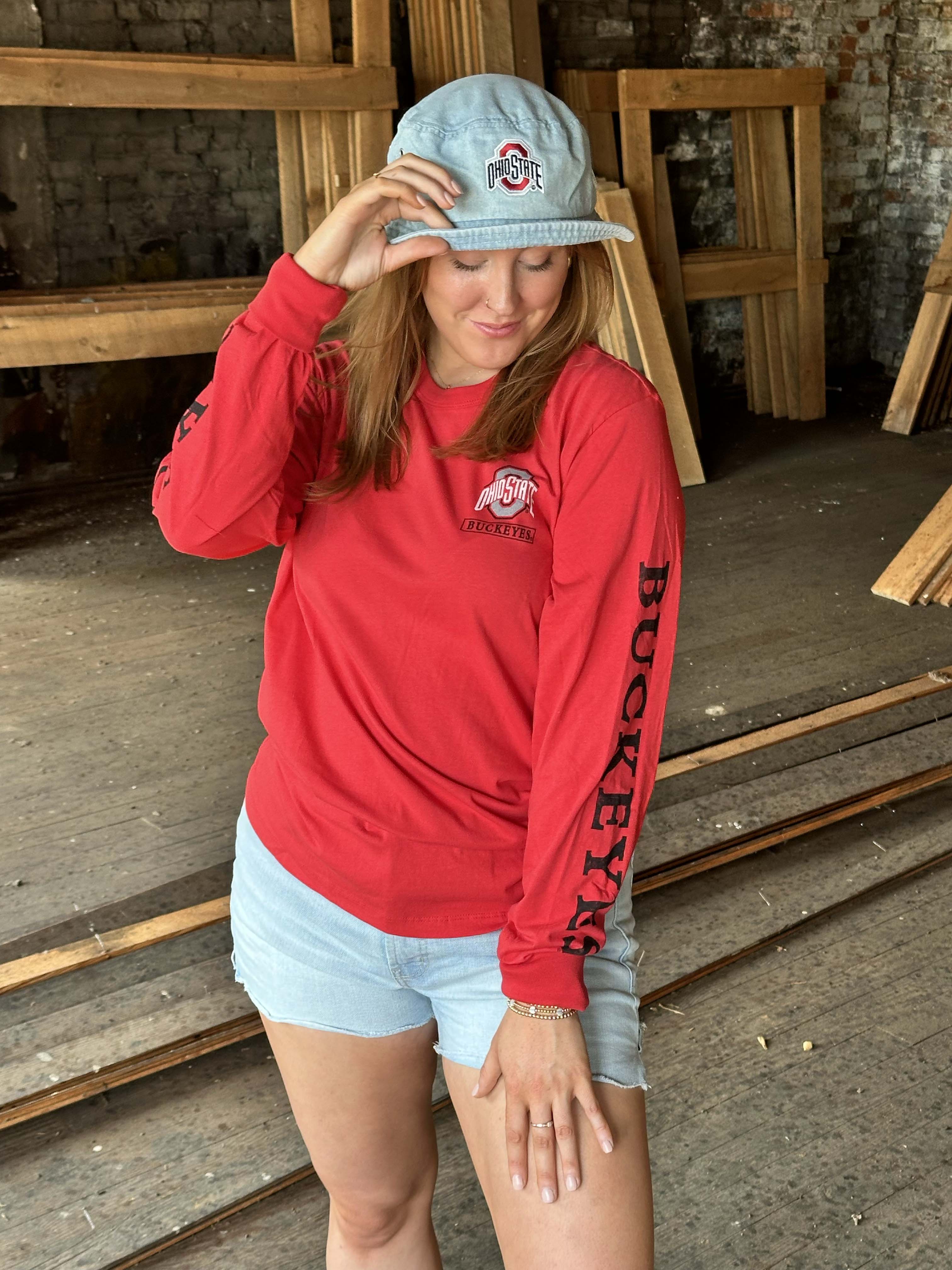 A woman wearing a bright red long-sleeve Ohio State Buckeyes sweatshirt, light blue denim shorts, and a light blue baseball cap with the Ohio State logo, standing in a rustic wood-paneled room.