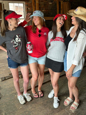 Four women laughing together in a rustic indoor setting, wearing Ohio State Buckeyes themed apparel including a gray graphic t-shirt, a red long-sleeve shirt, a gray knit 'Buckeyes' sweater, and a white button-up shirt.