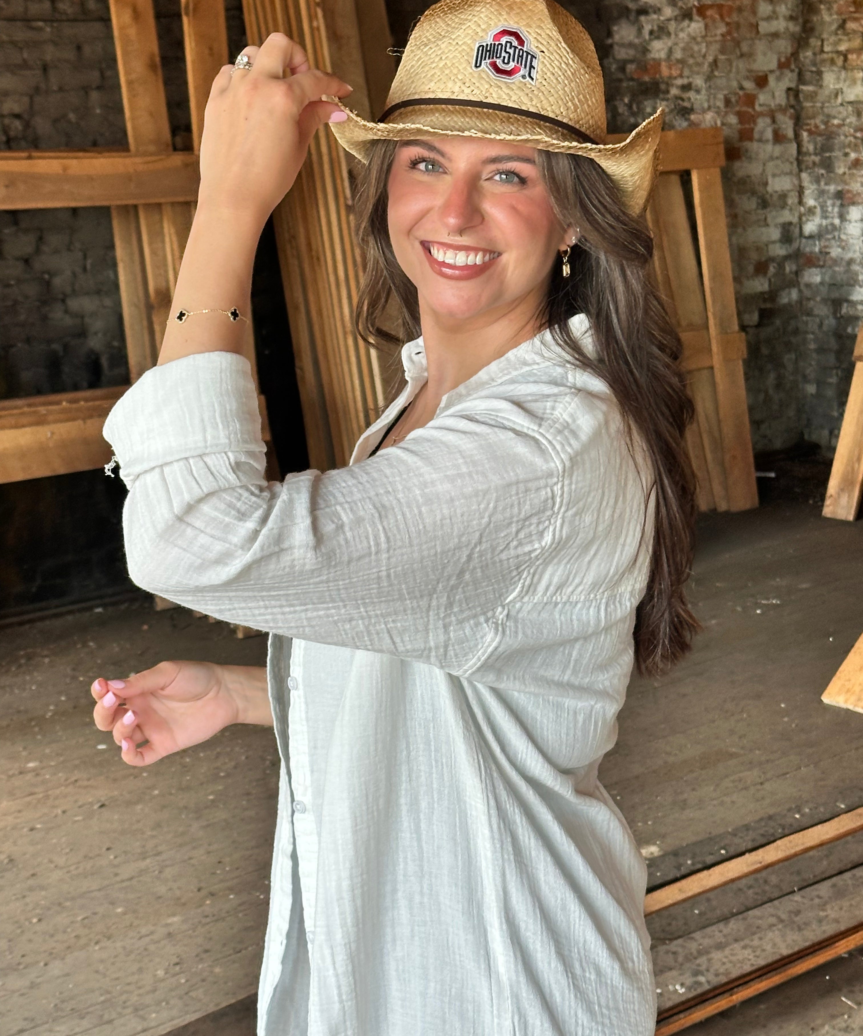 A woman smiling and wearing a straw cowboy hat with an Ohio State Buckeyes logo, paired with a light gray long-sleeve button-up shirt, standing in a rustic indoor setting with wooden beams and brick walls.