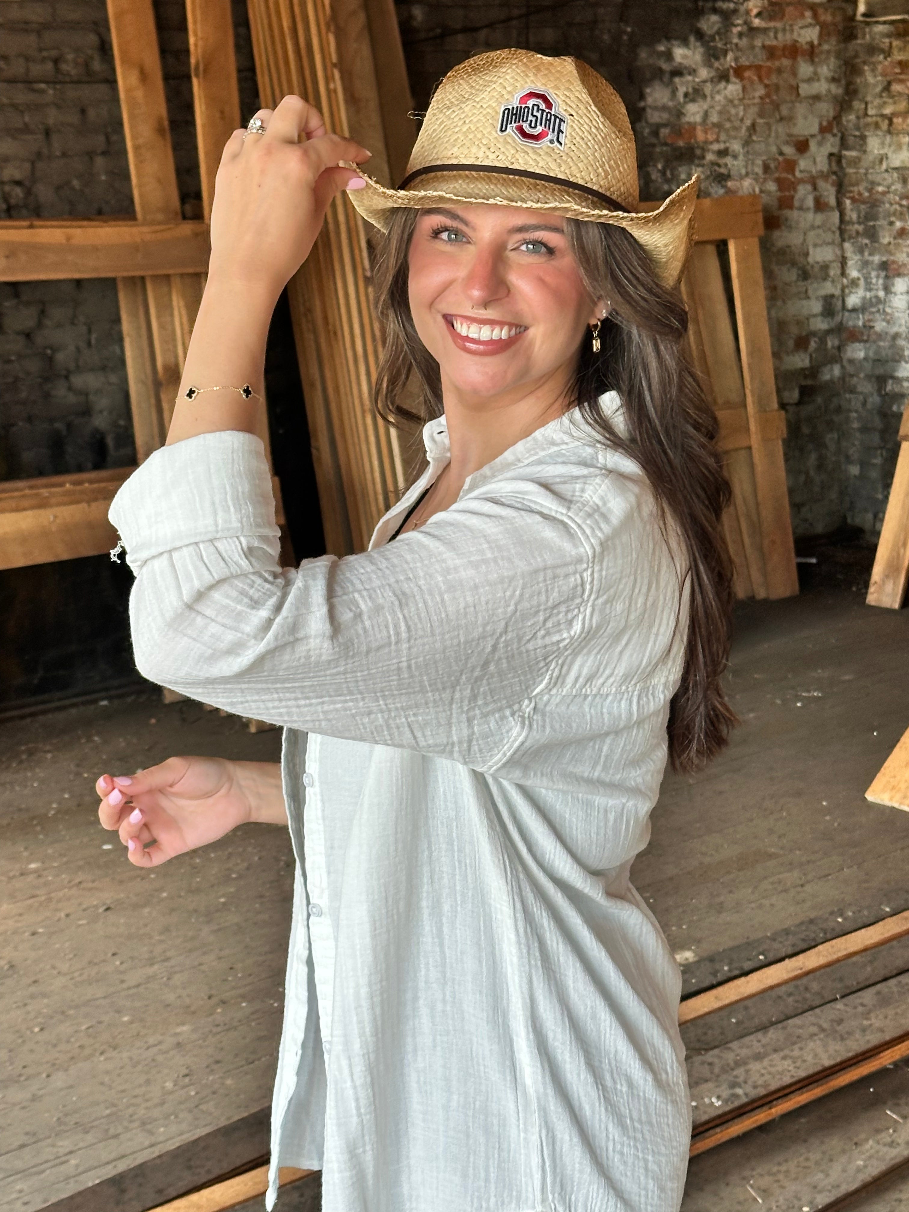 A woman smiling and wearing a straw cowboy hat with an Ohio State Buckeyes logo, paired with a light gray long-sleeve button-up shirt, standing in a rustic indoor setting with wooden beams and brick walls.