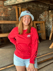 A smiling woman wearing a red long-sleeve t-shirt with a 'Pittsford' logo, a light blue bucket hat, and denim shorts, standing in a rustic workshop with brick and wood walls.