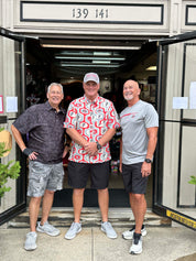 Three men standing together in front of an open storefront doorway, wearing casual summer clothing. The storefront has a beige exterior with the address 139 141 above the entrance.