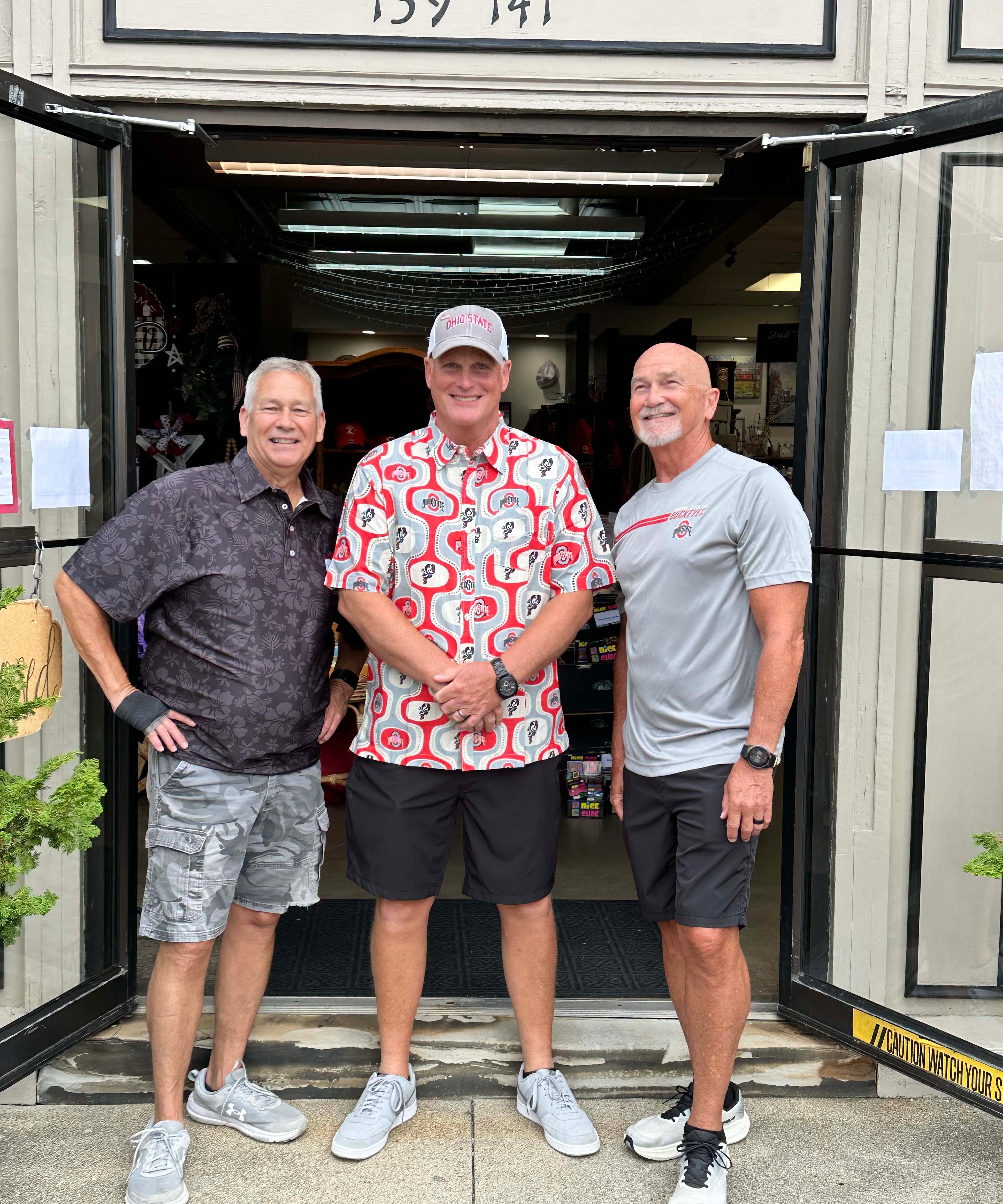 Three men standing together in front of an open storefront doorway, wearing casual summer clothing. The storefront has a beige exterior with the address 139 141 above the entrance.