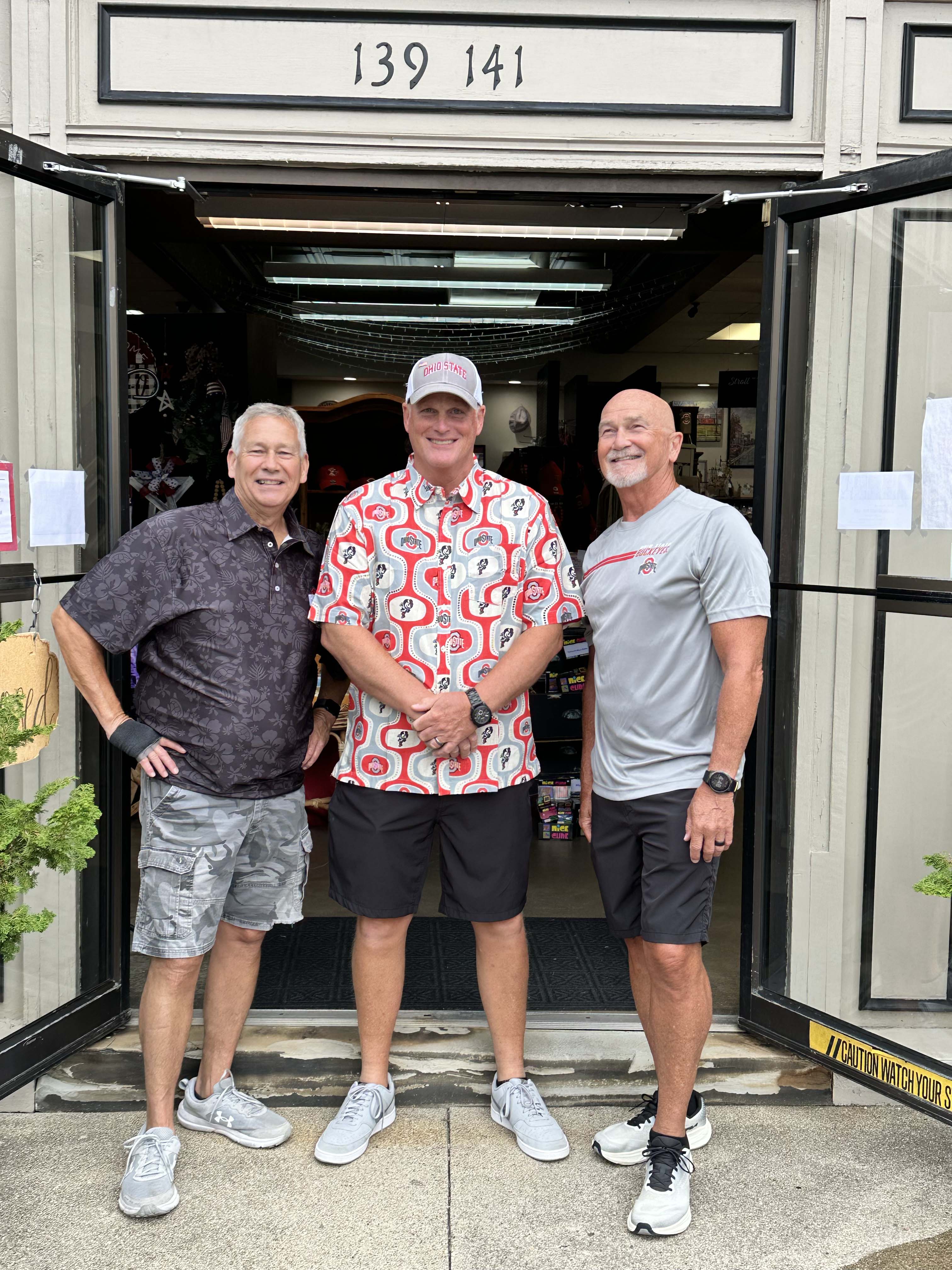 Three men standing together in front of an open storefront doorway, wearing casual summer clothing. The storefront has a beige exterior with the address 139 141 above the entrance.