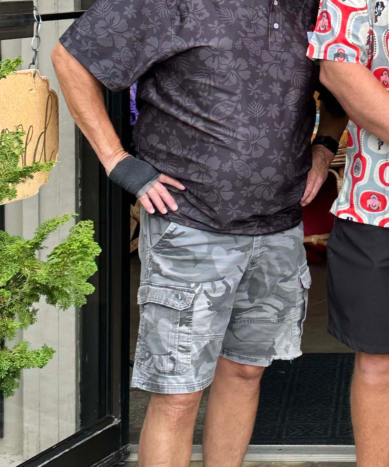 An older man standing outdoors in front of a storefront doorway, wearing a dark patterned short-sleeve polo shirt, gray cargo shorts, and light gray sneakers. Another person is partially visible on the right edge.