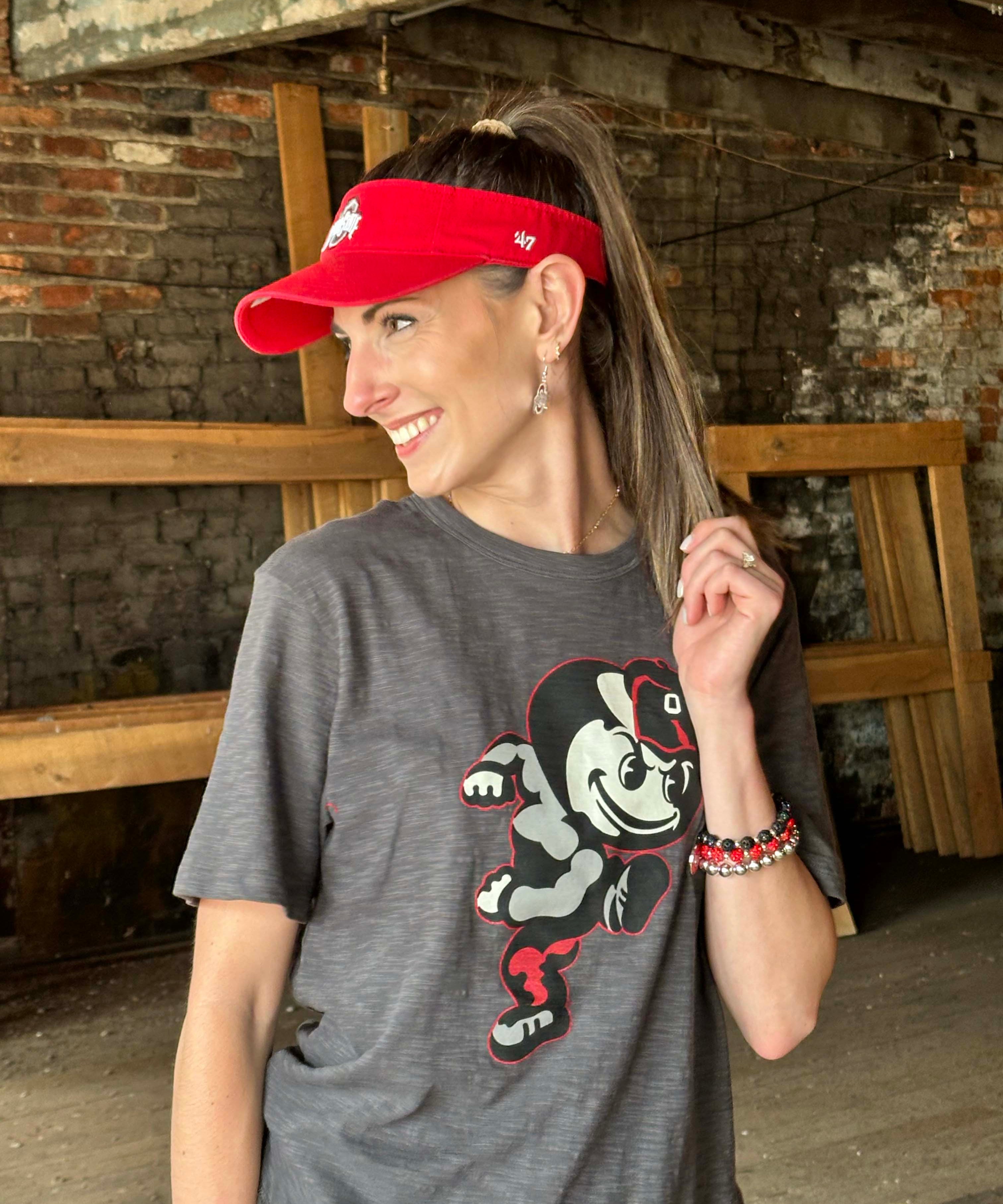 A woman wearing a dark gray graphic t-shirt with a cartoon mascot and a red visor, standing in a rustic brick and wood interior.