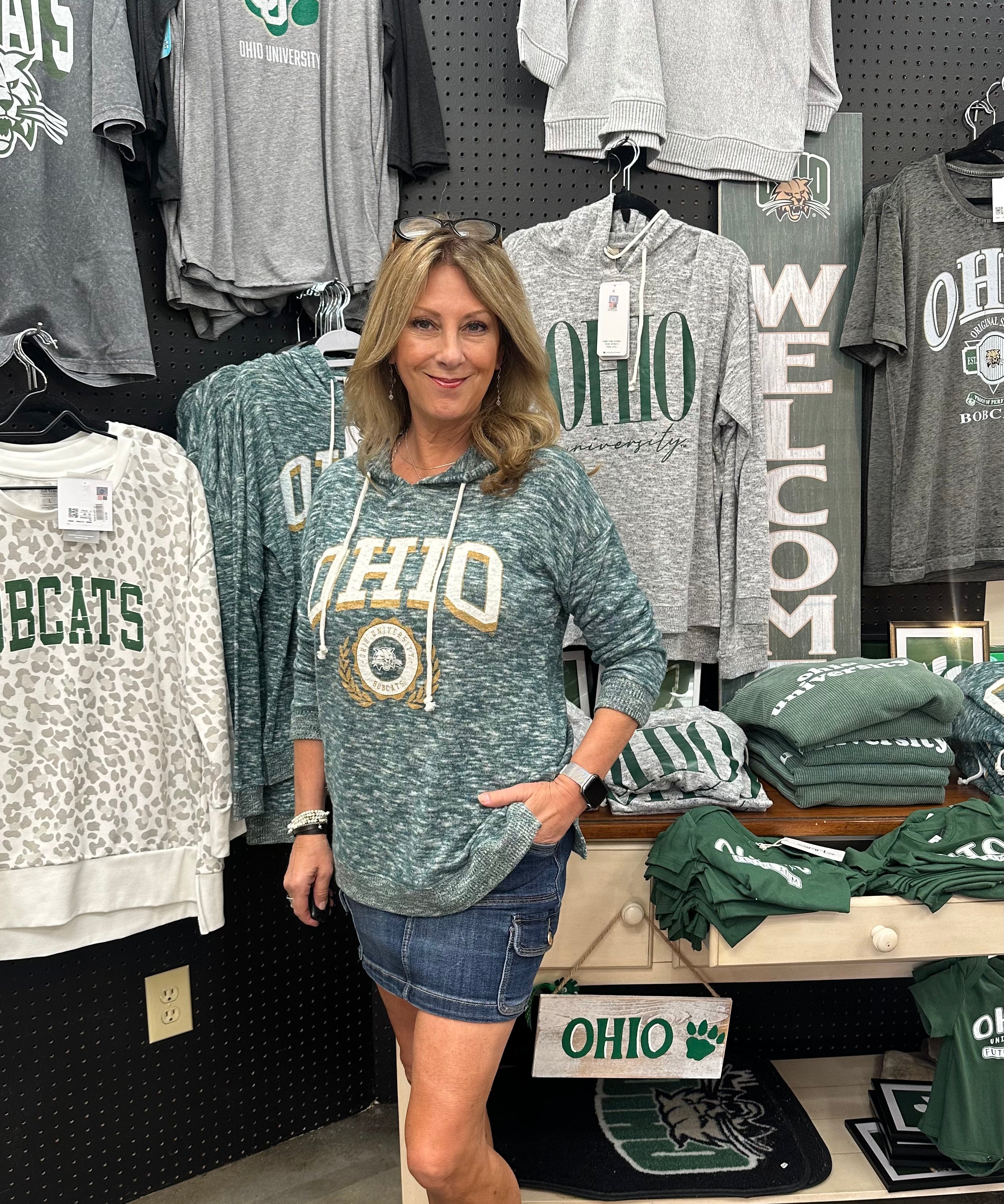 Woman wearing a green 'OHIO' sweatshirt in a store with various clothing items on display.
