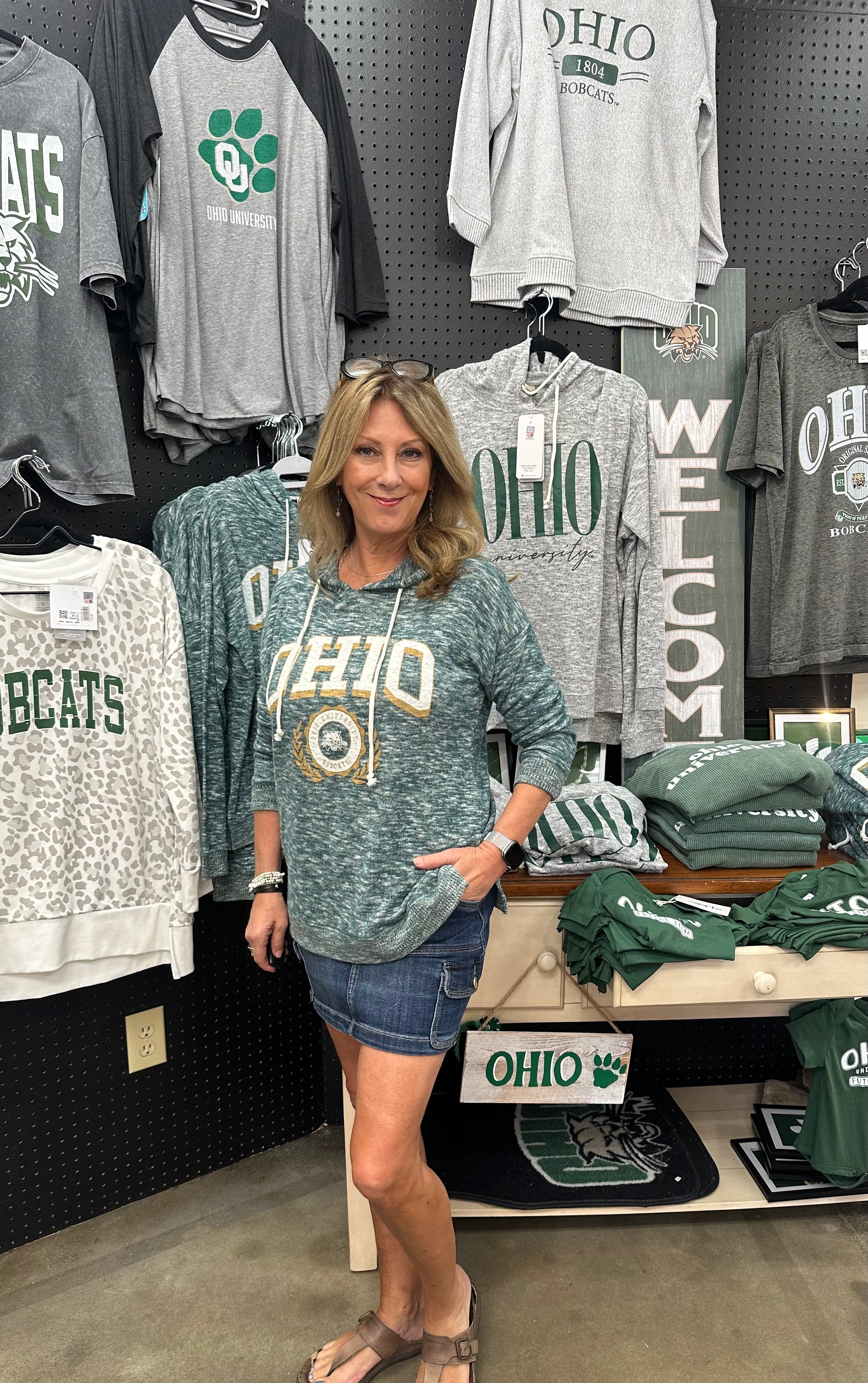 Woman wearing a green 'OHIO' sweatshirt in a store with various clothing items on display.