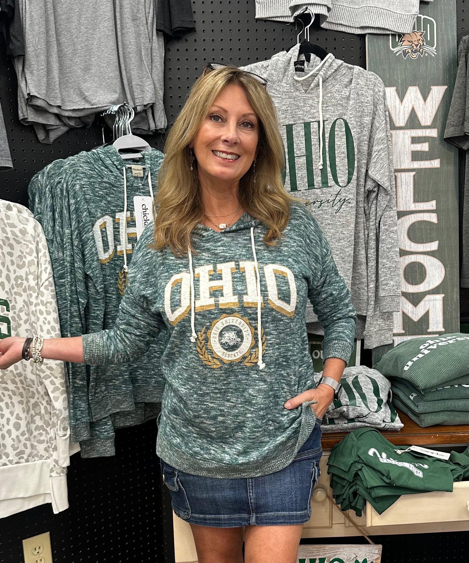 Woman wearing a green 'OHIO' sweatshirt in a store setting with various clothing items around.