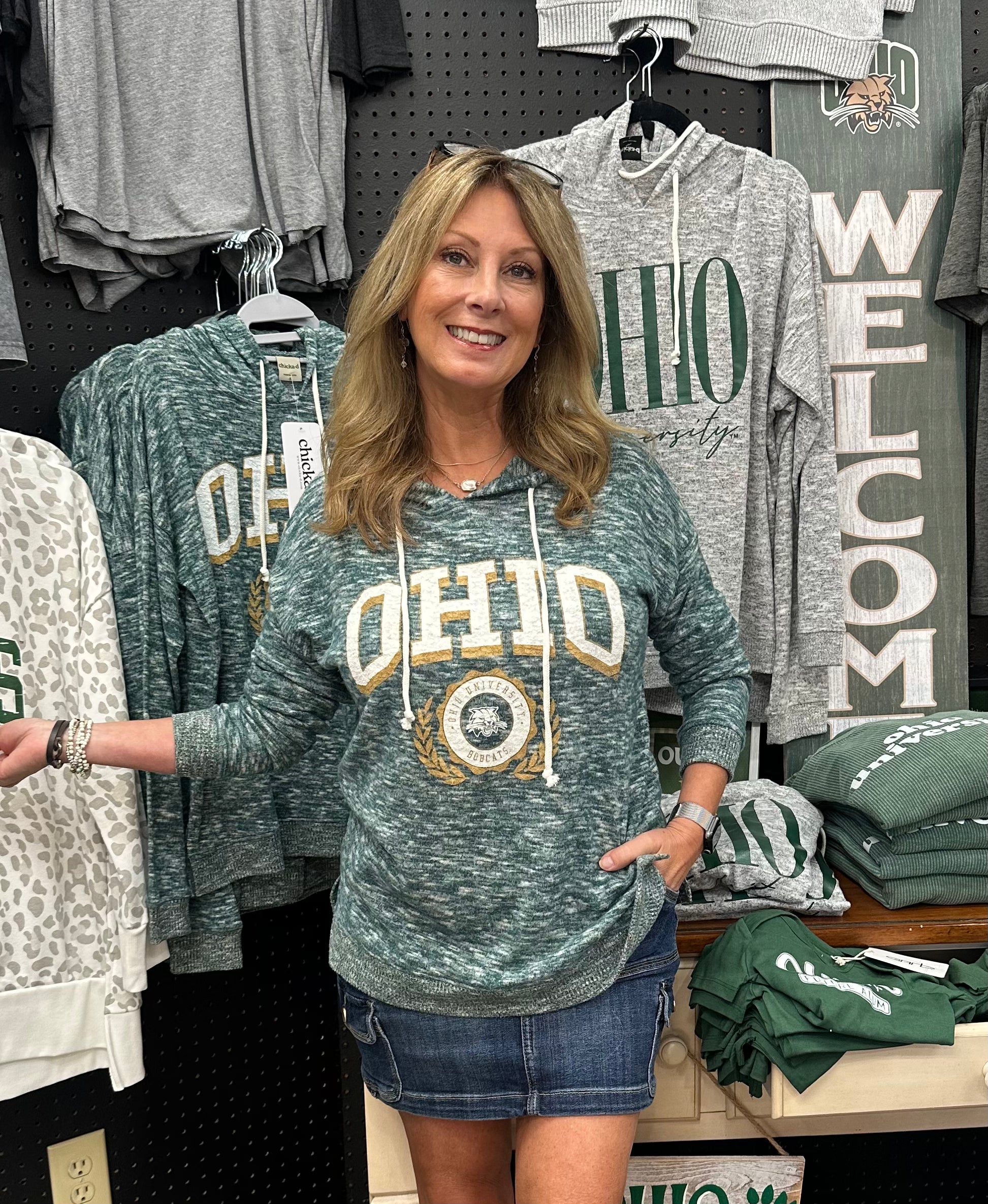 Woman wearing a green 'OHIO' sweatshirt in a store setting with various clothing items around.