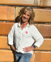 A smiling blonde woman wearing a white long-sleeve button-up shirt with a small red logo on the chest, blue jeans, and a red necklace, standing against a tan brick wall.