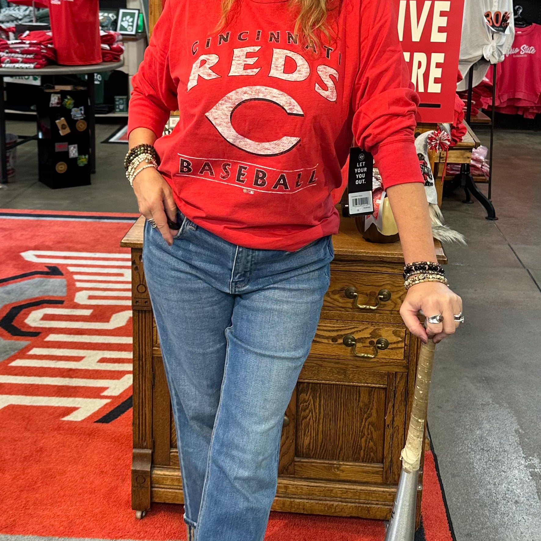 Woman in a red sports jersey and cap holding a baseball bat in a store with sports-themed decor.