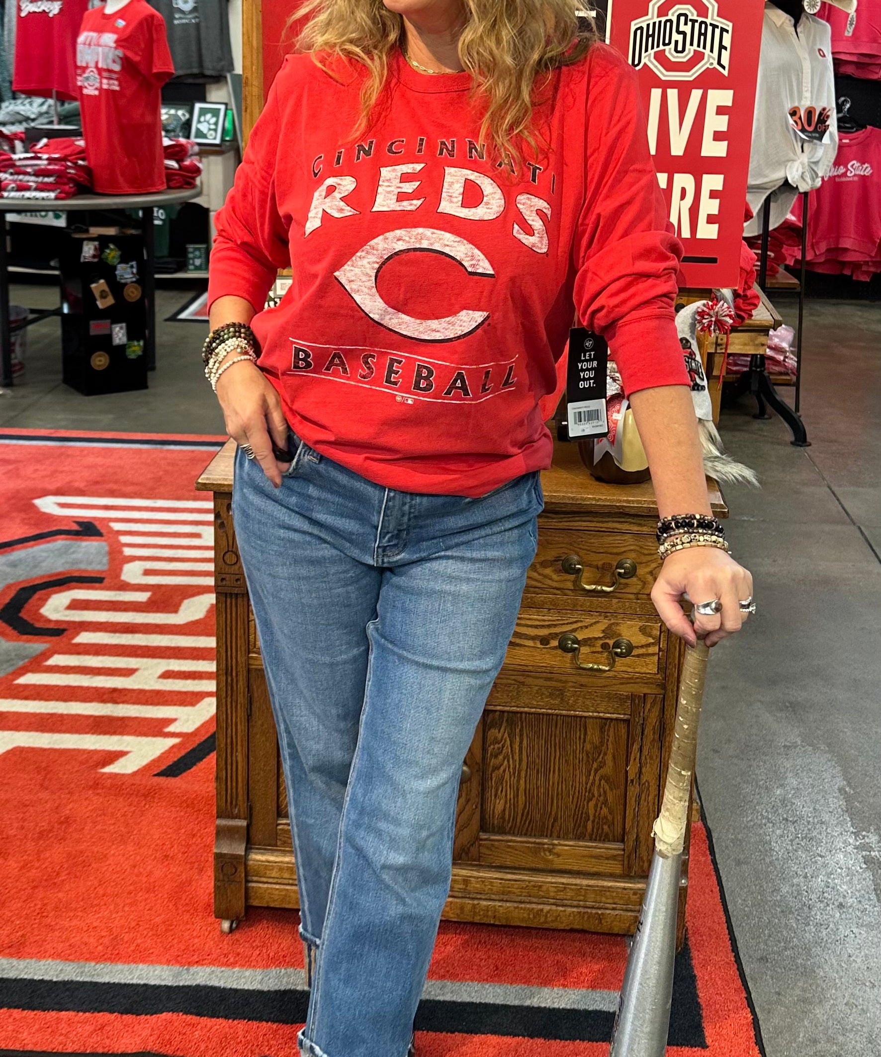 Woman in a red sports jersey and cap holding a baseball bat in a store with sports-themed decor.