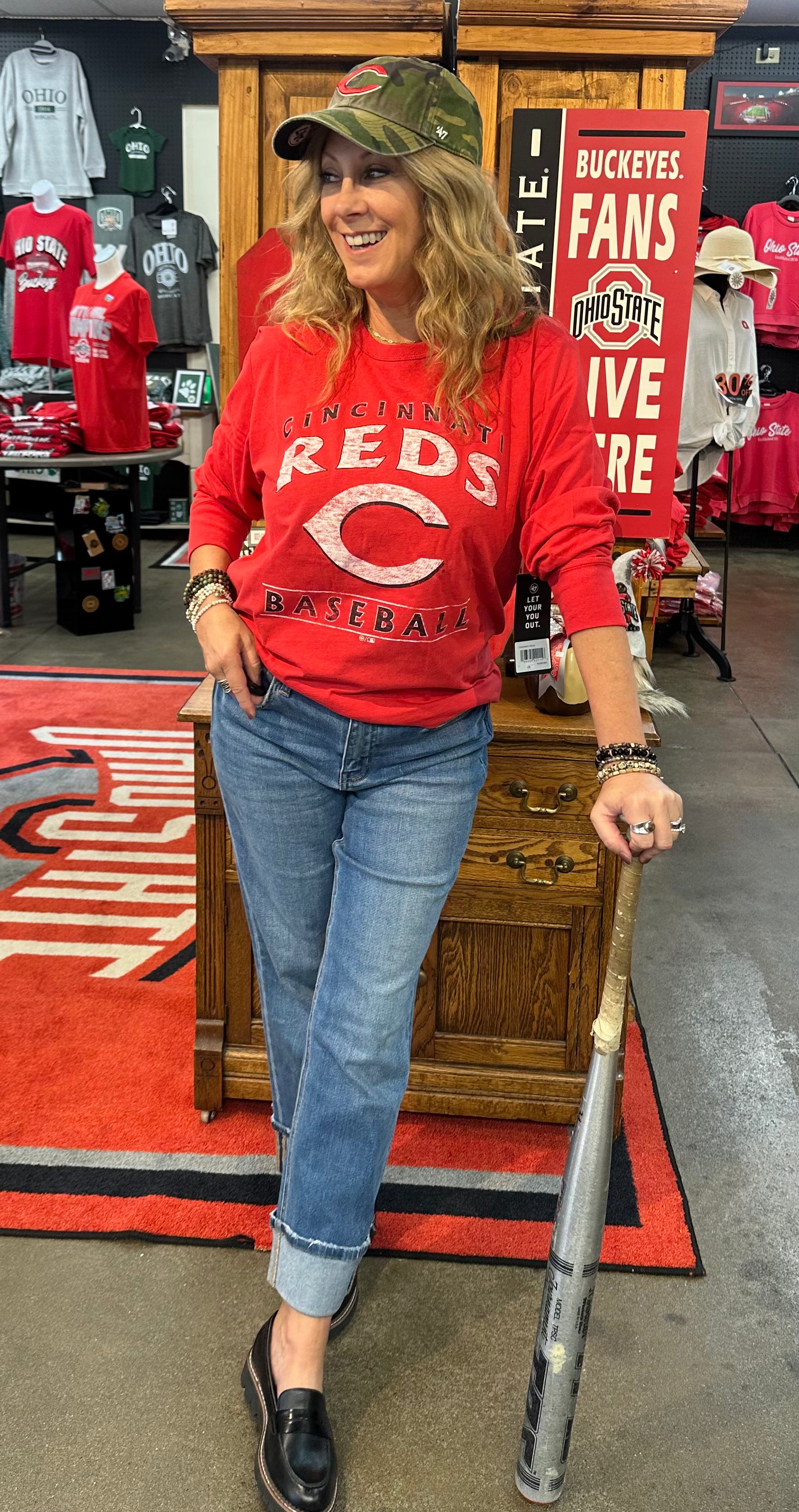 Woman in a red sports jersey and cap holding a baseball bat in a store with sports-themed decor.