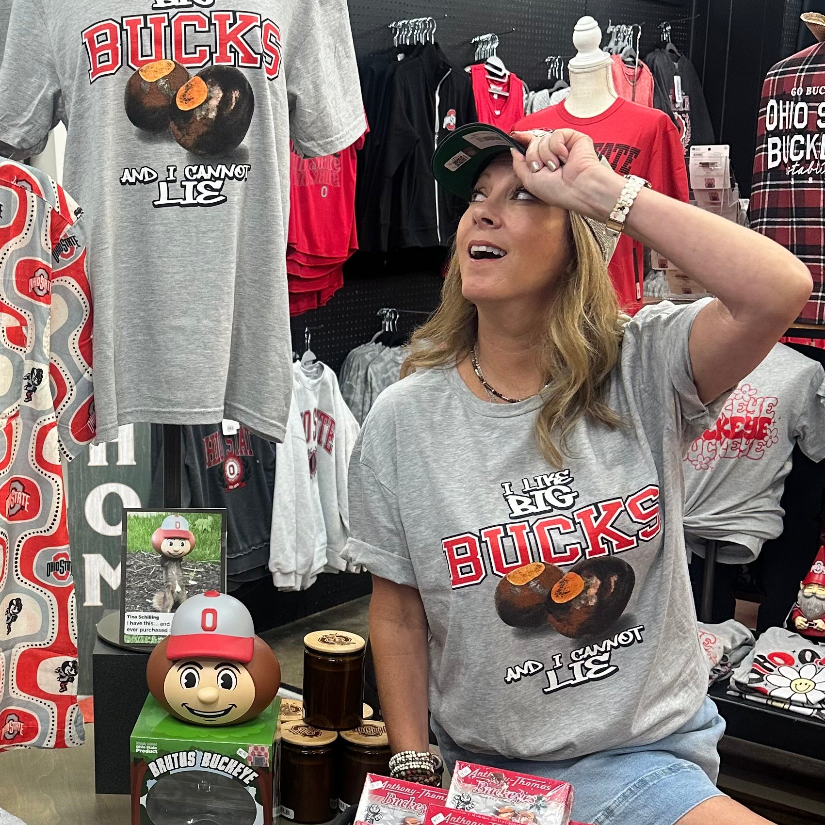 A woman wearing a gray Ohio Buckeyes t-shirt sits in a retail store next to a mannequin displaying a matching gray t-shirt with a cowboy hat. The gray t-shirt features 'The Ohio Big Buckies' and 'We're Making Lies' printed in red and orange.